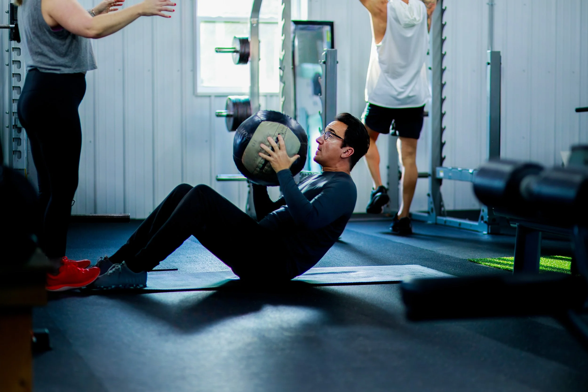 A man doing sit-ups with a medicine ball while a woman stands nearby in a gym, with workout equipment and other people exercising in the background.