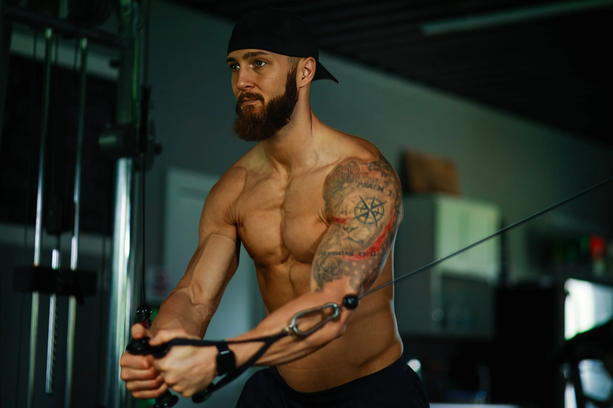 A shirtless man with tattoos and a beard, wearing a black cap backward, is exercising with a cable machine in a gym.