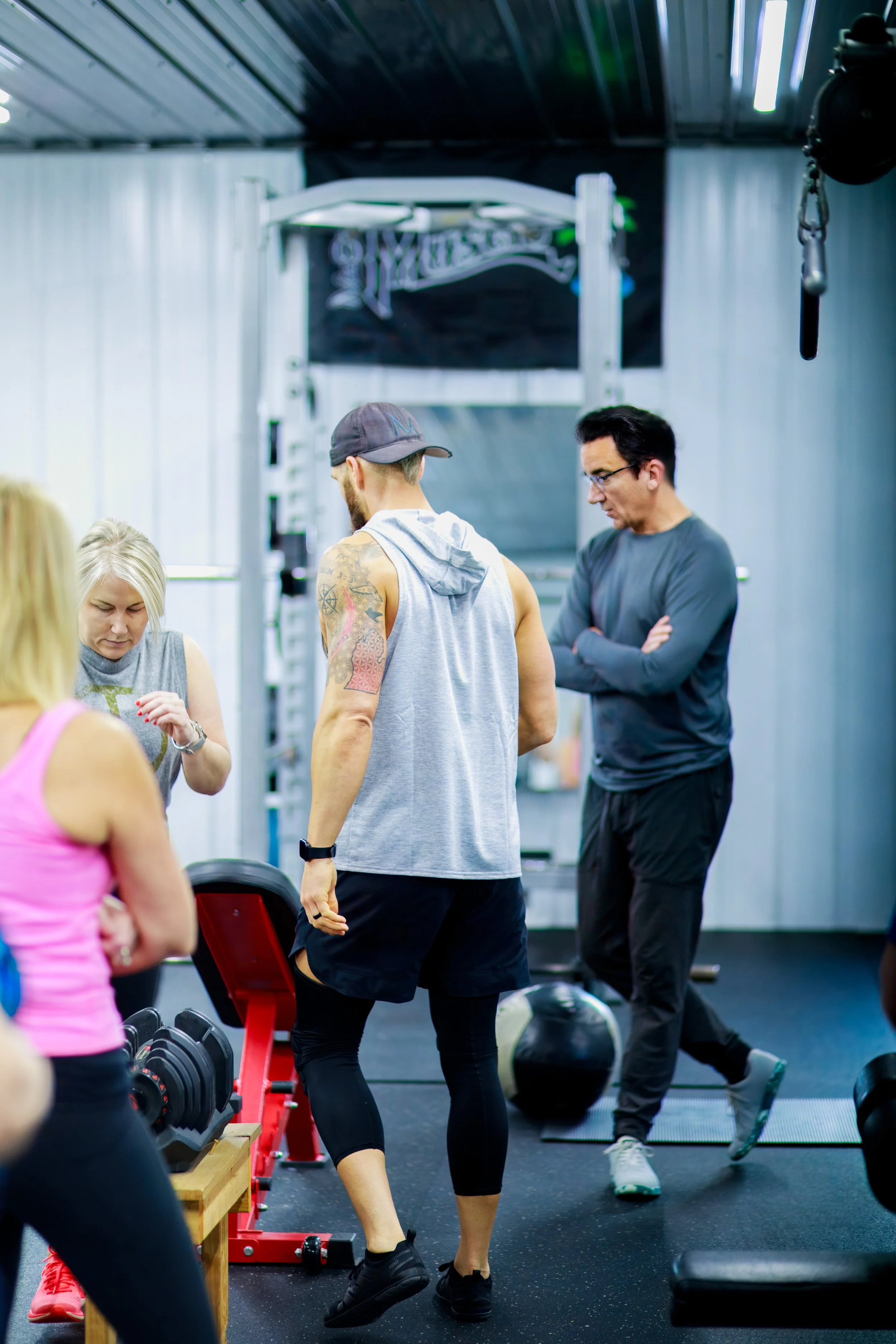 People working out in a gym, with one person lifting weights and others observing or preparing to exercise.
