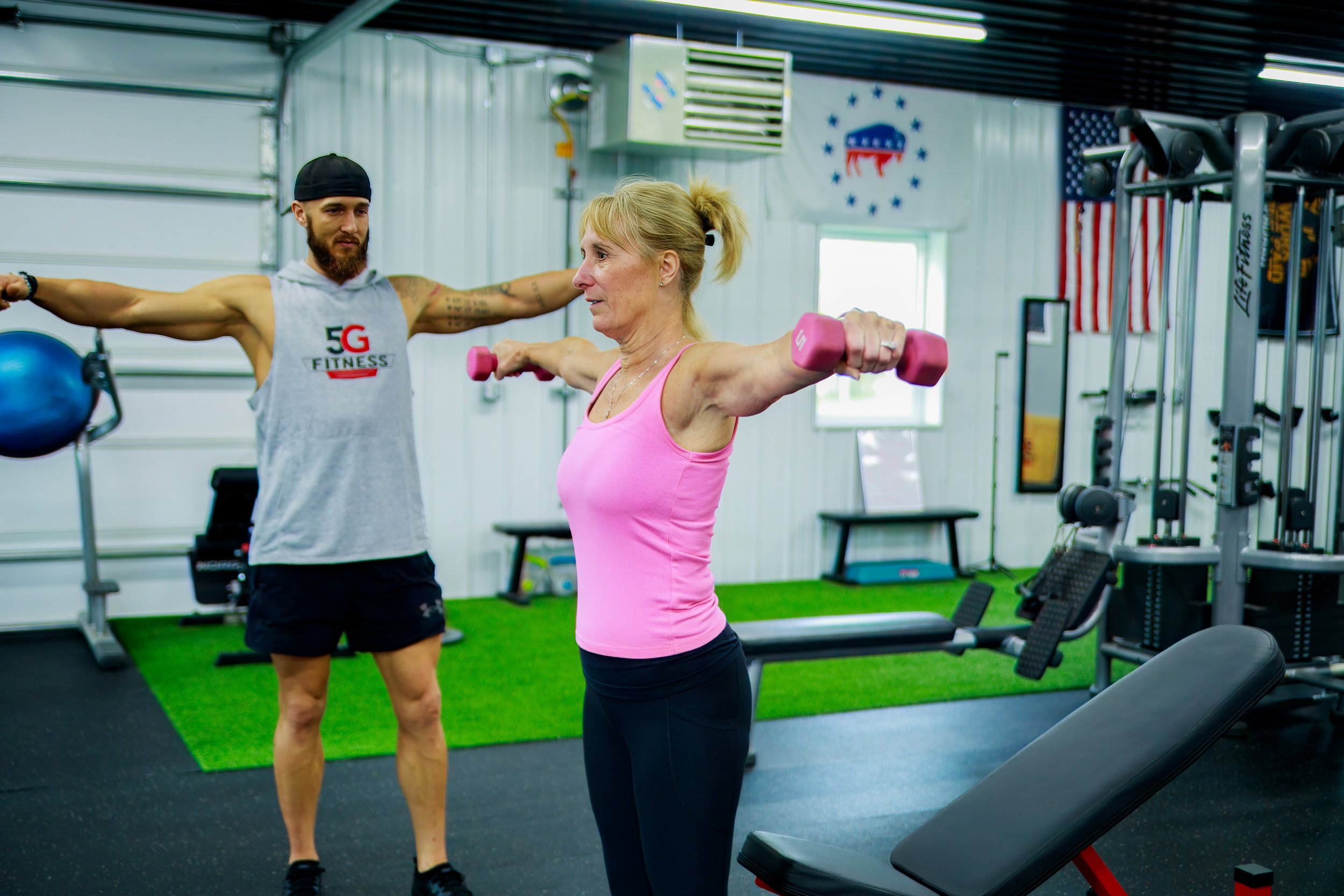 A woman in a pink tank top and black workout pants lifting pink dumbbells in a gym, assisted by a trainer standing beside her with arms outstretched. The gym has American flags and gym equipment visible in the background.