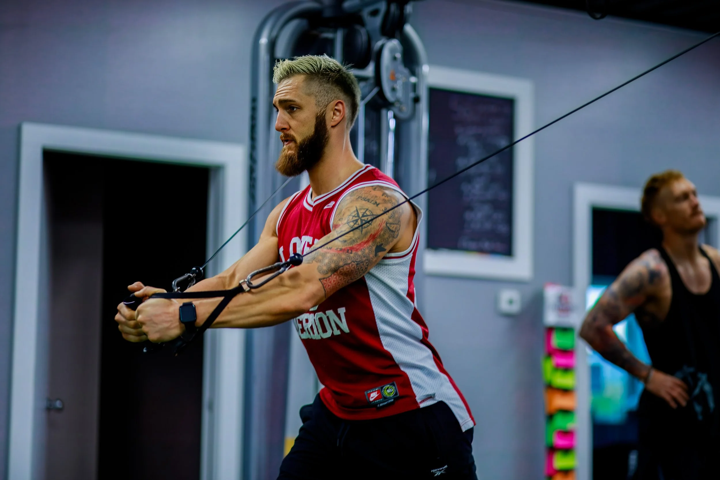 Man with a beard and tattoos exercising on cable machine at gym, wearing a red and white sports jersey.