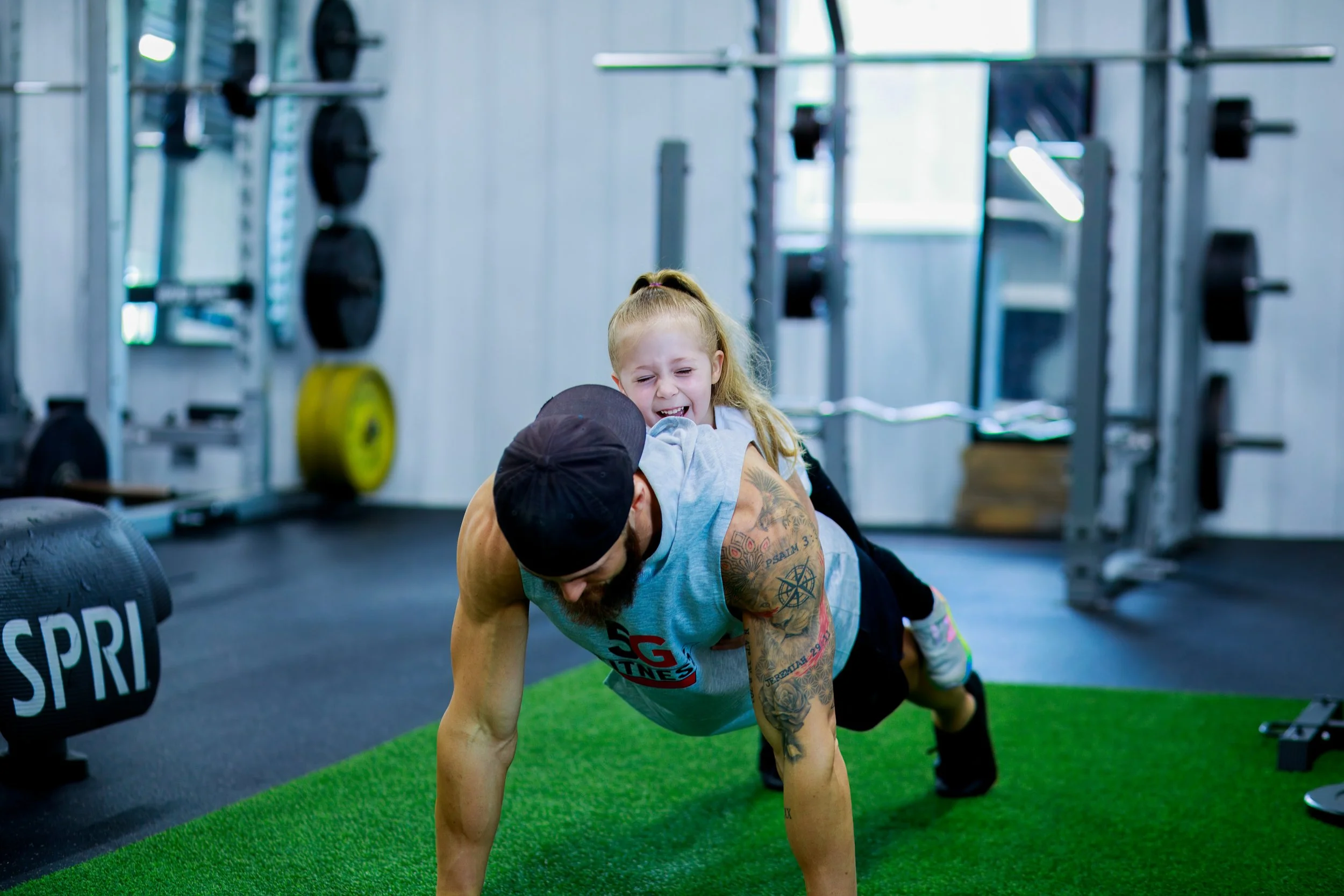 A man doing push-ups on a green mat while a young girl playfully sits on his back in a gym with fitness equipment in the background.