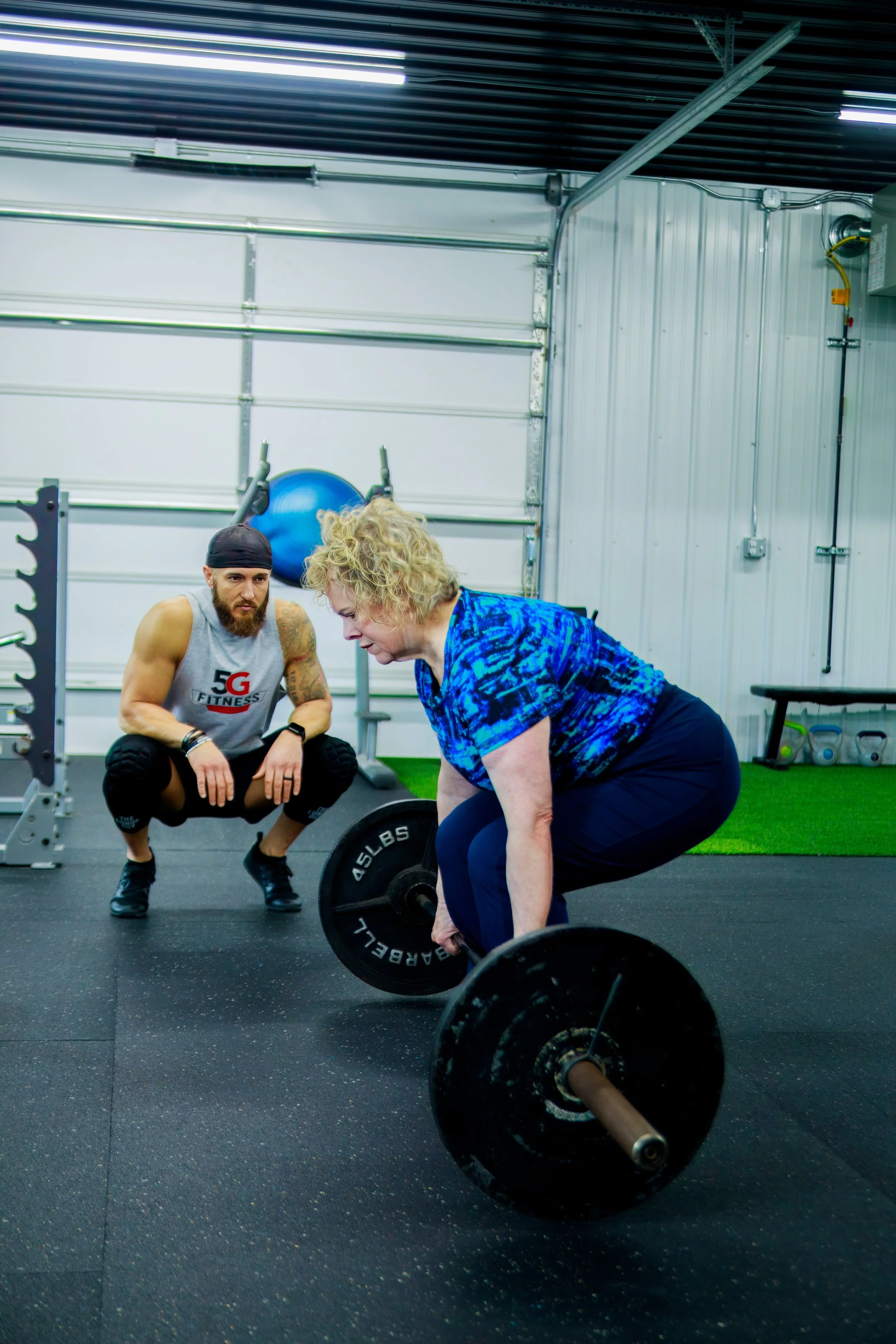 An older woman doing a deadlift exercise with a barbell in a gym, guided by a trainer.
