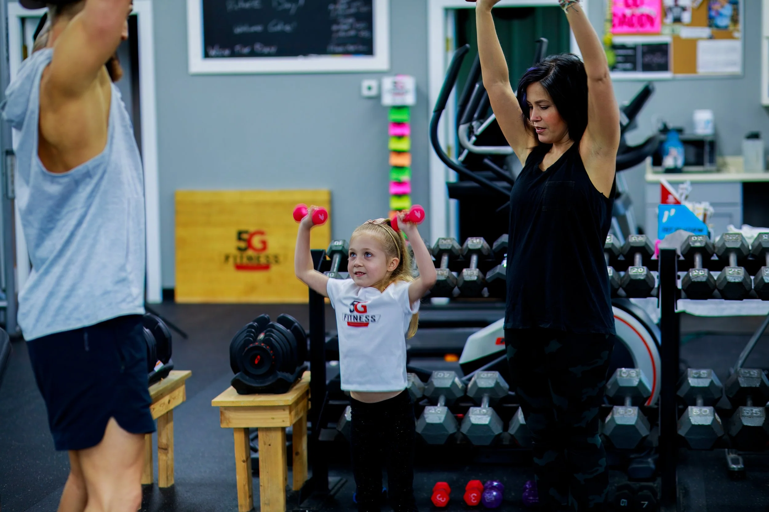 A young girl lifting pink dumbbells in a gym, guided by an instructor, with another woman standing nearby. The gym has various weights, exercise equipment, and colorful signs in the background.