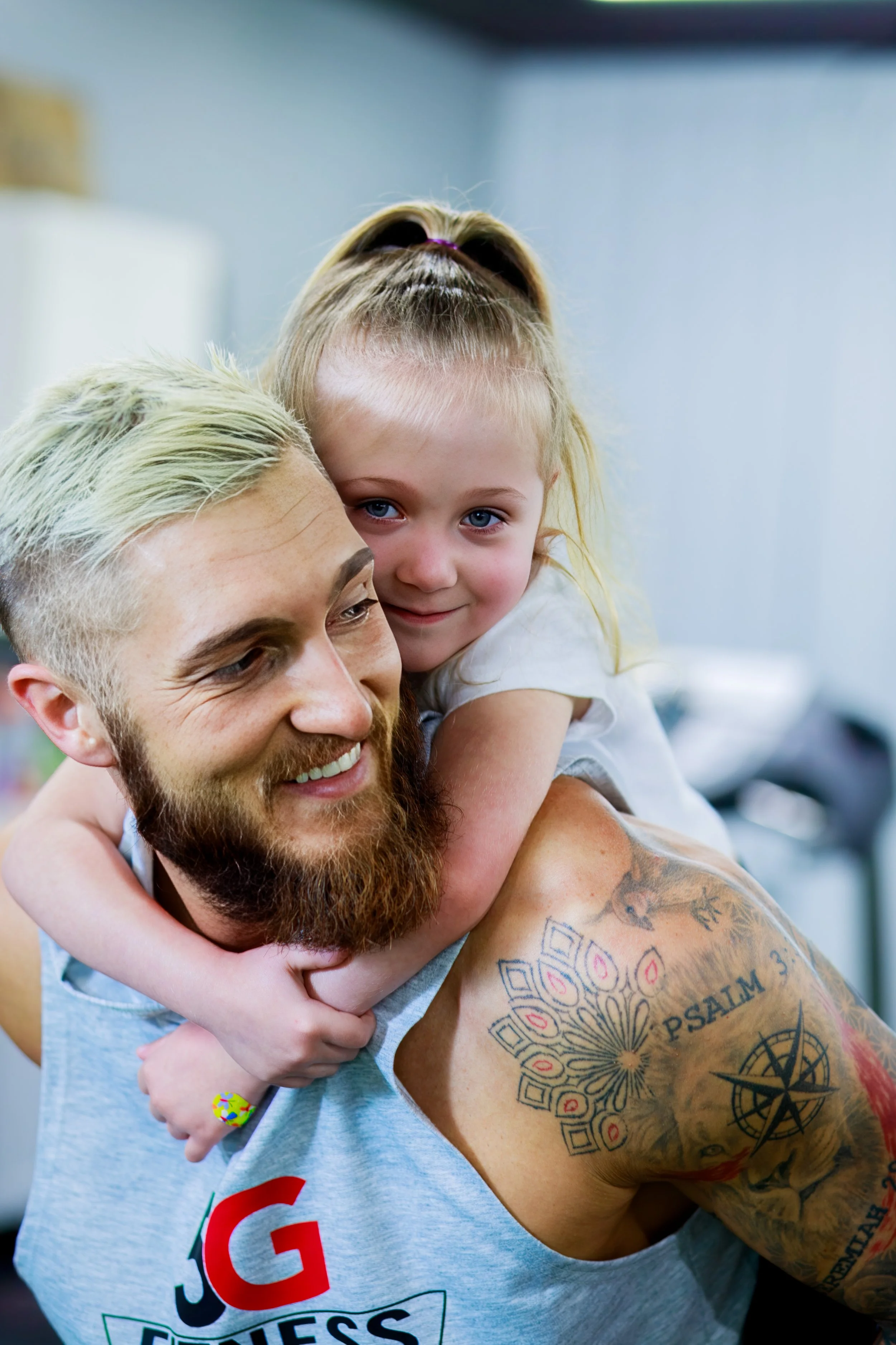 A man with a beard and tattoos smiling as a young girl with blonde hair in a ponytail hugs him from behind.