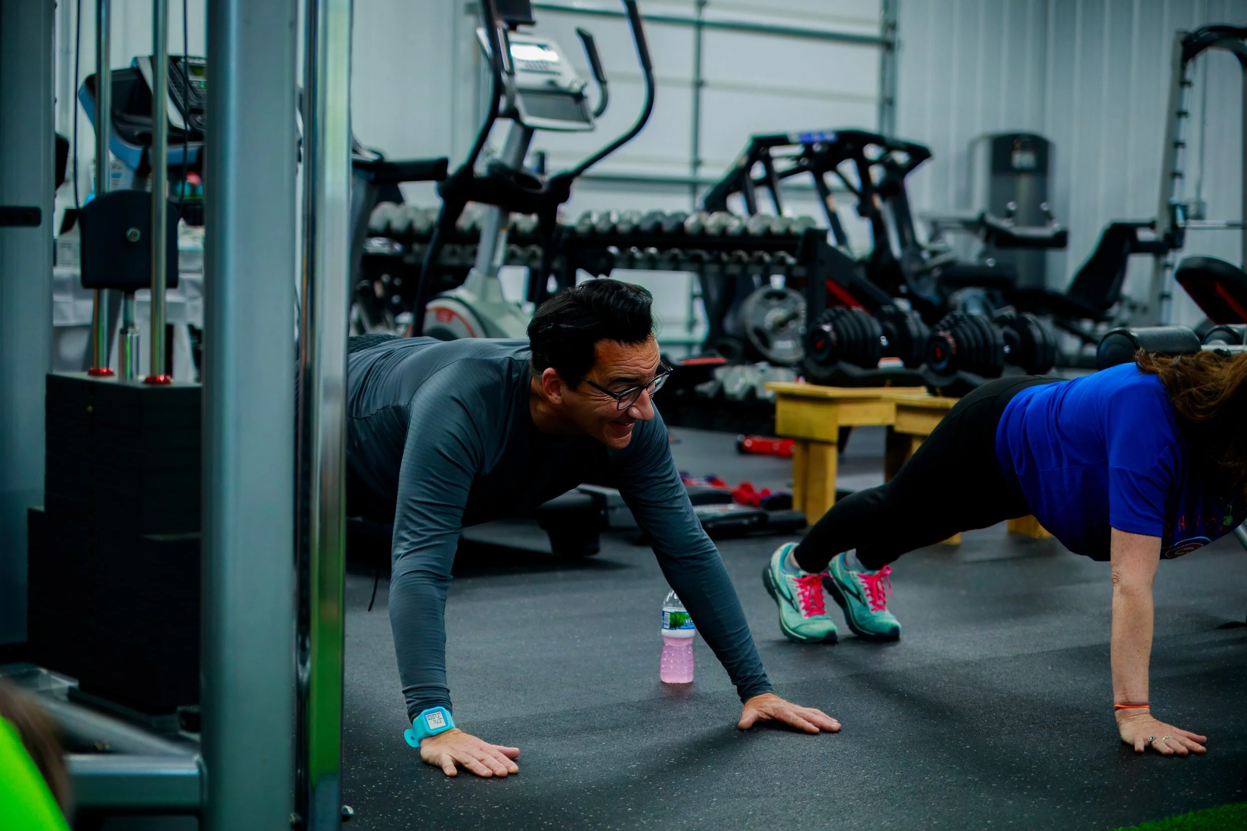 Two people doing push-ups on the gym floor surrounded by exercise equipment.
