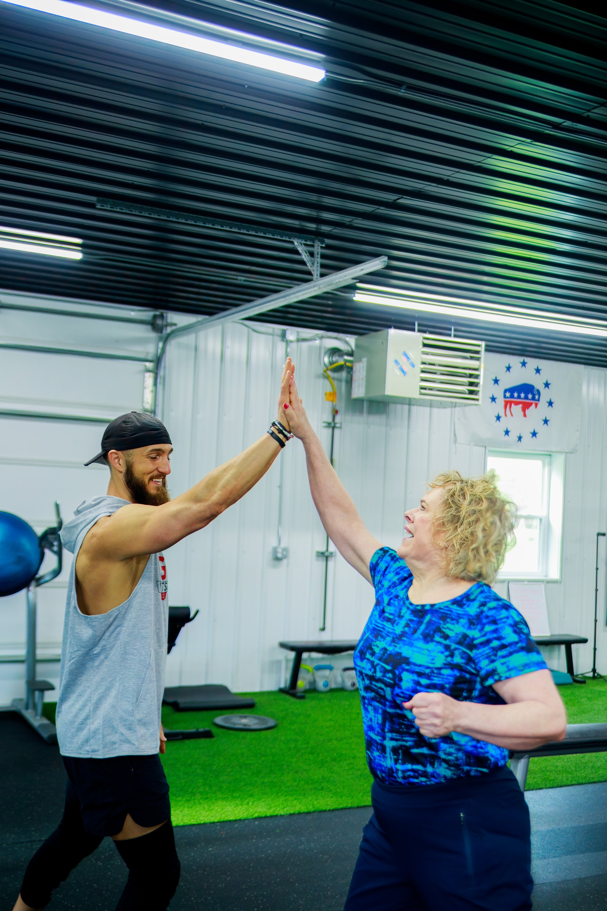 A man and a woman high-five each other inside a gym with fitness equipment and an American political campaign logo on the wall.
