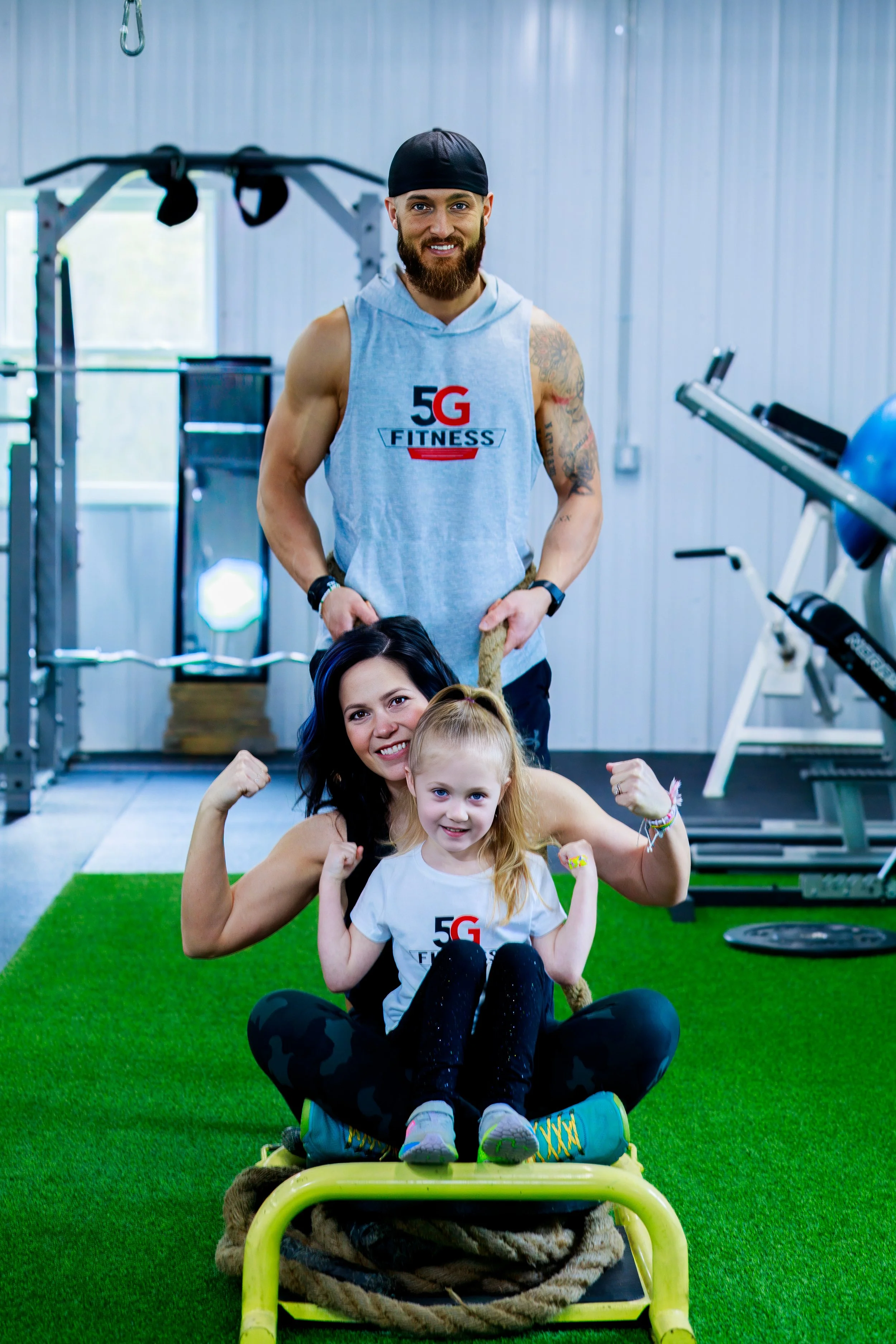 A family of three at the gym: a fit man with a beard stands behind a woman and a young girl, all smiling and flexing their muscles, with gym equipment and a green turf floor in the background.