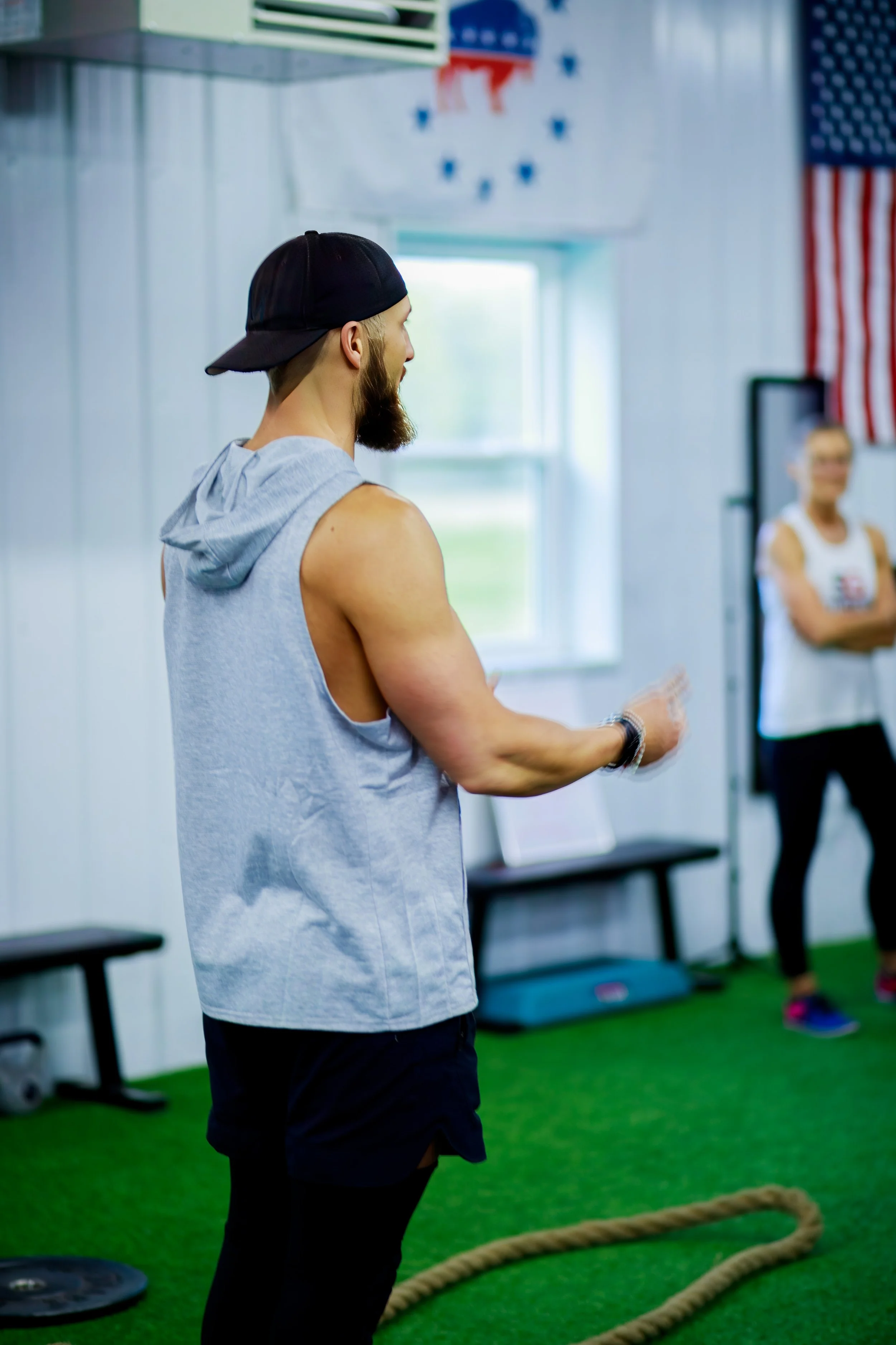 A muscular man with a beard, wearing a sleeveless gray hoodie and a backward black cap, standing in a gym with green turf flooring. Whiteted woman with glasses, white tank top, and black leggings stands in the background with arms crossed. American f