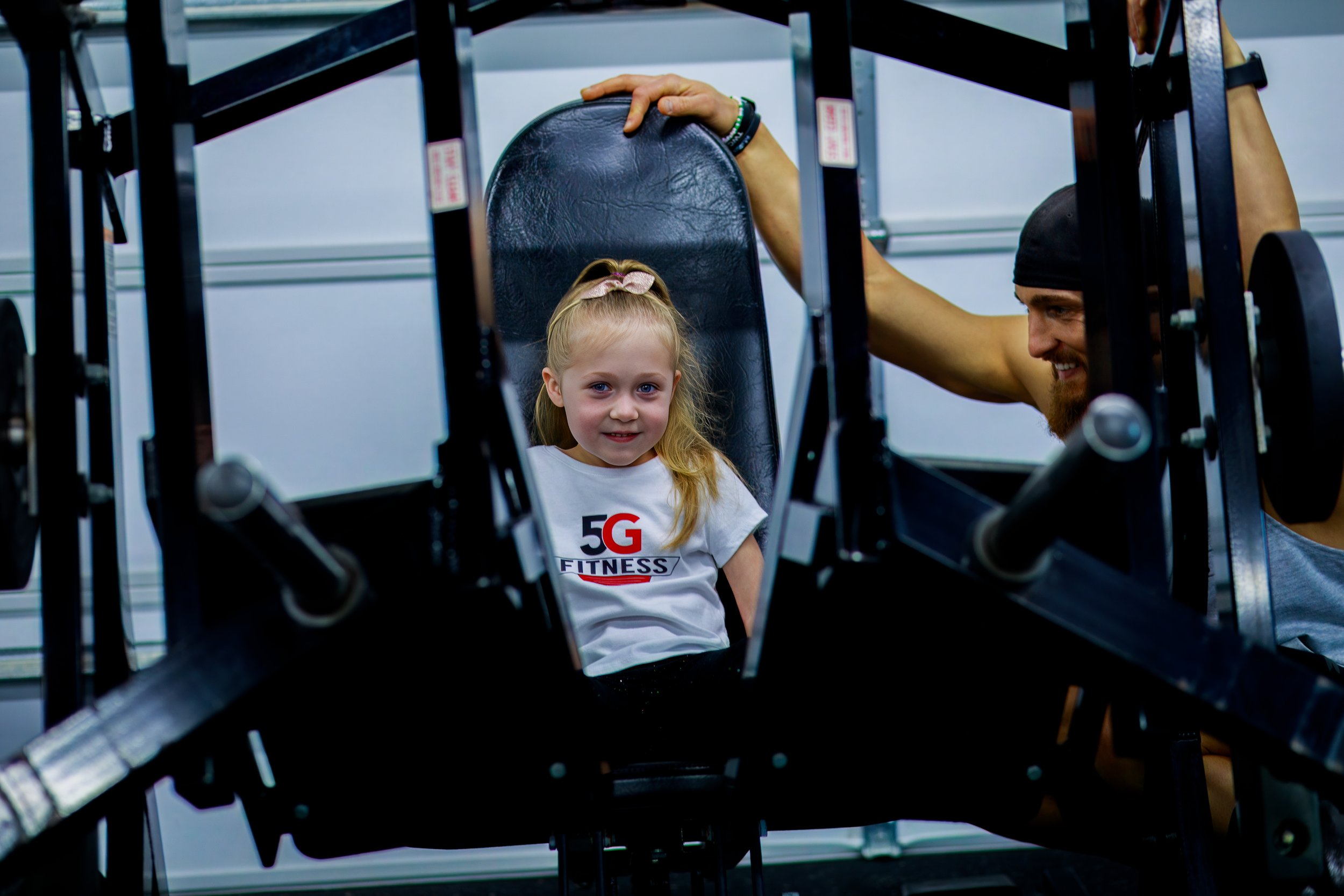 A young girl with blonde hair sitting on exercise equipment in a gym, wearing a white T-shirt with '5G FITNESS' printed on it. A man with a beard and a black head covering is visible smiling behind her.