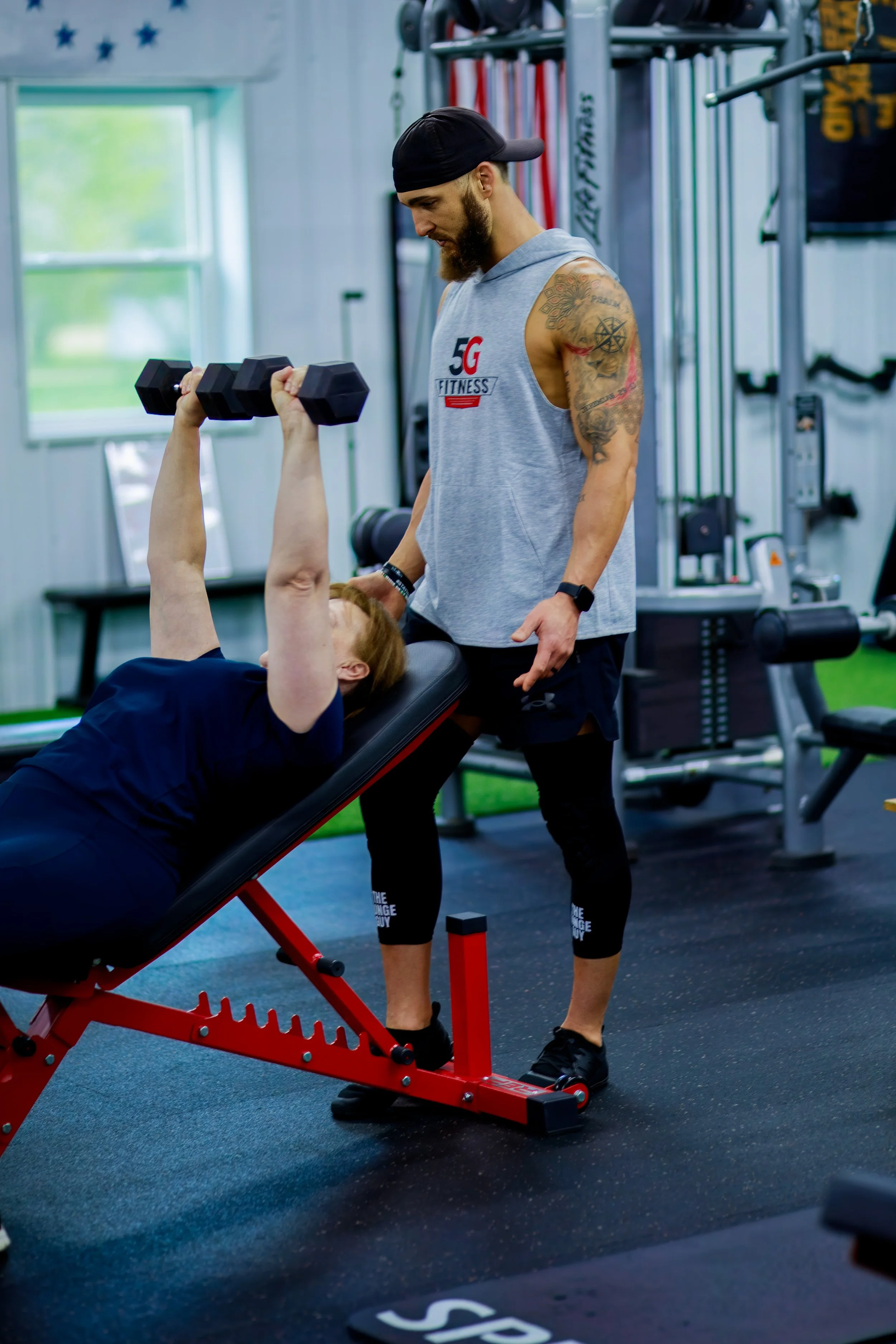 A woman lying on a workout bench lifting dumbbells while a trainer stands beside her in a gym.