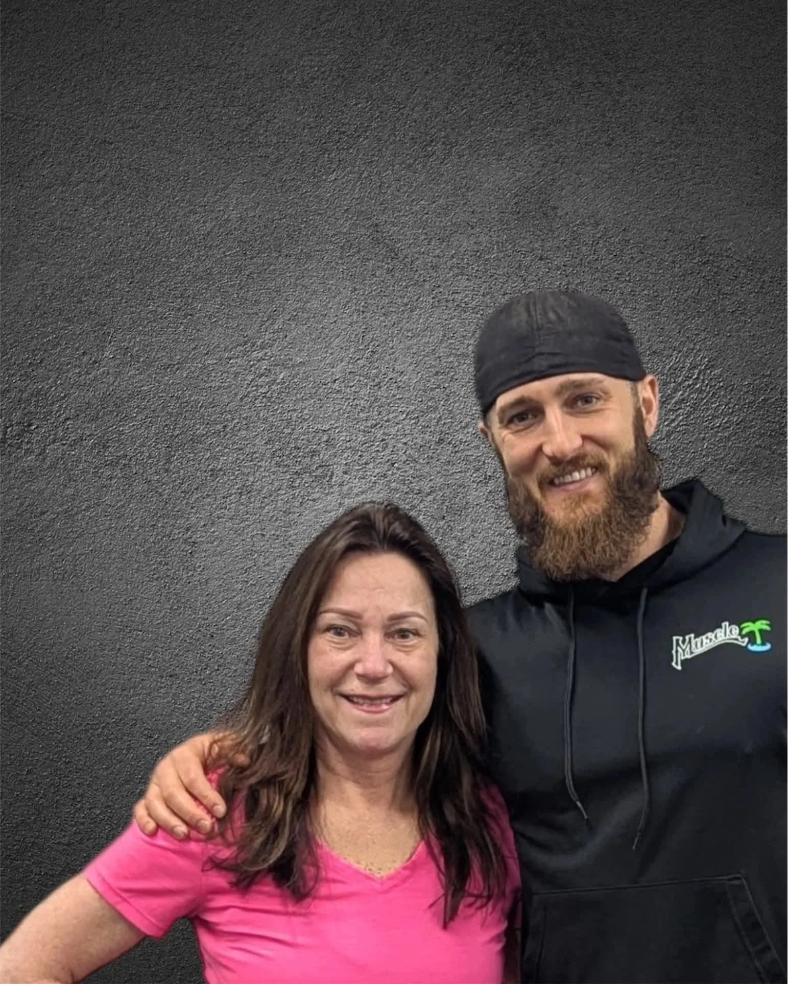 A woman with brown hair wearing a pink t-shirt and a man with a beard and black head covering wearing a black hoodie, standing together against a dark textured wall, smiling at the camera.