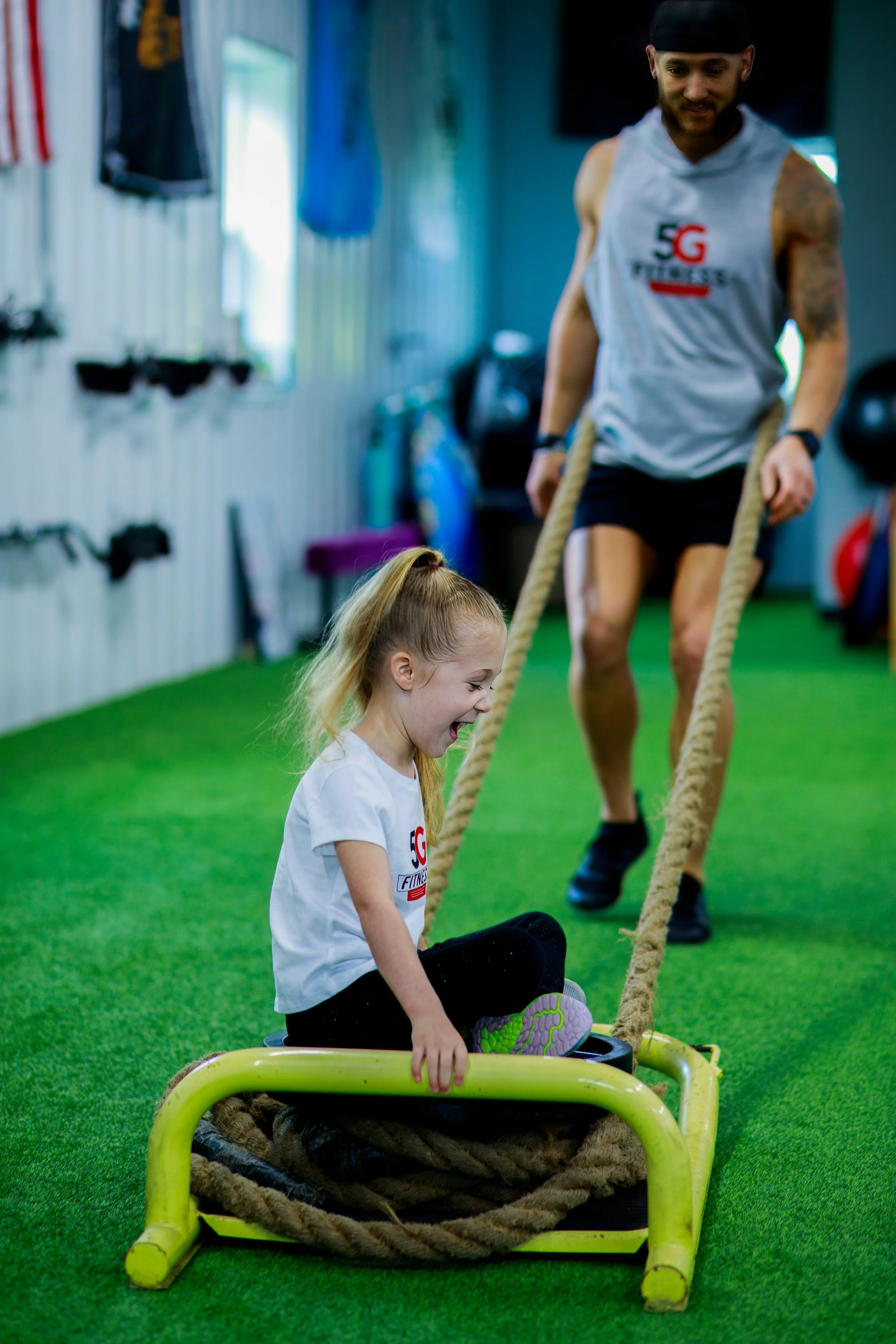 A young girl sitting in a sled, laughing, while a man pulls her using a rope. They are in a gym with green turf flooring, with workout equipment on the walls.