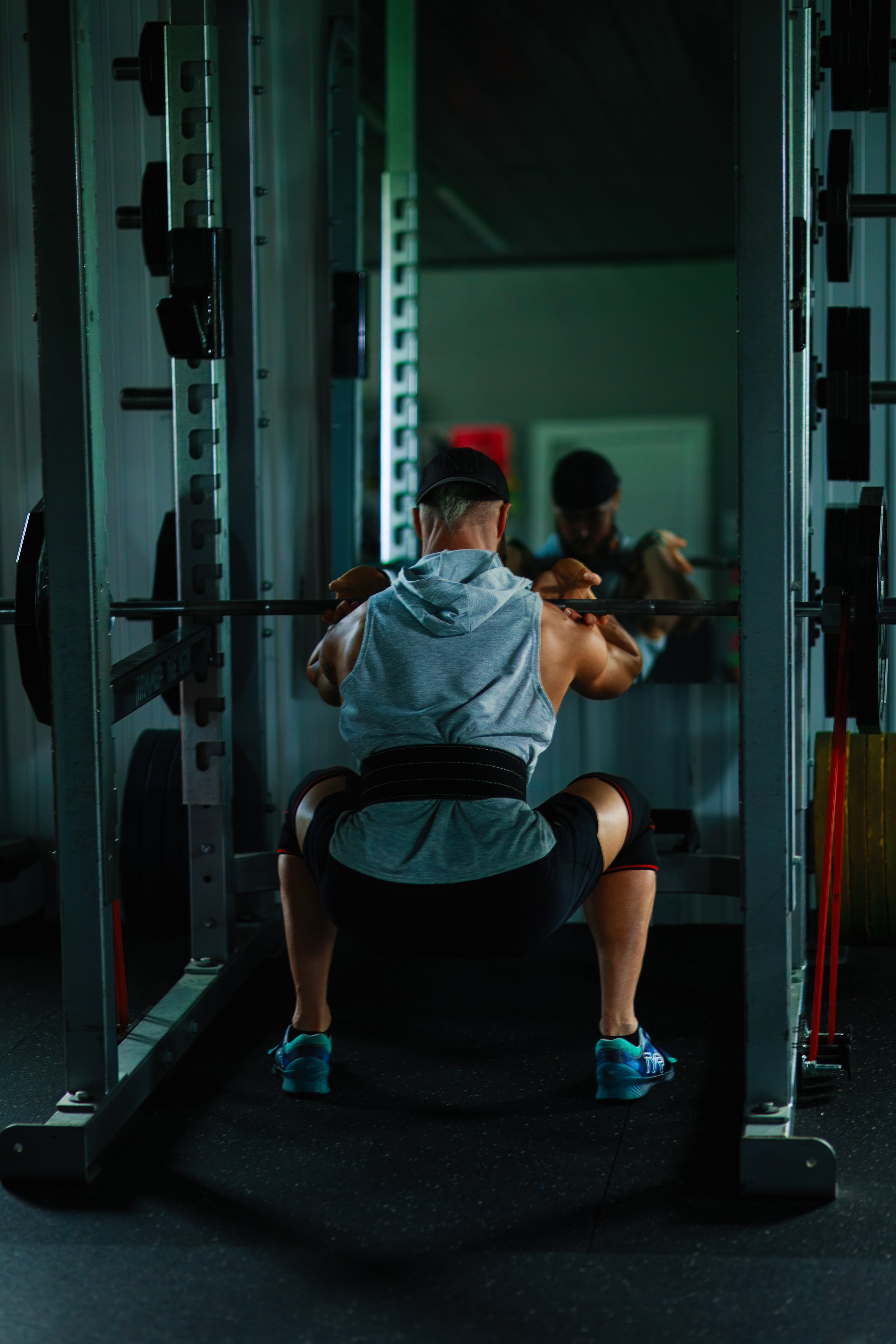 Man squatting with a barbell in a gym, reflected in a mirror, wearing a sleeveless hoodie, shorts, and sneakers.