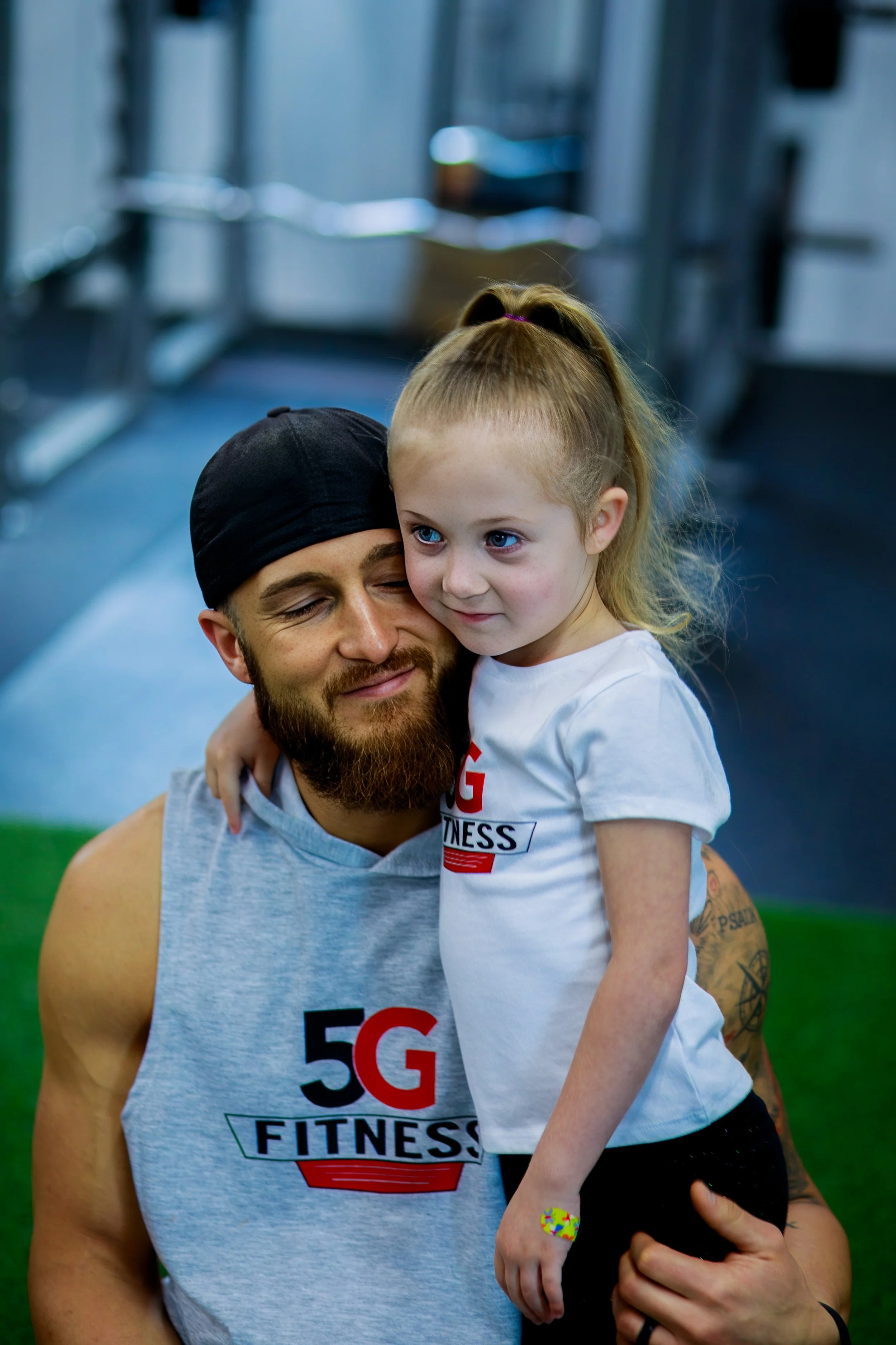 A bearded man wearing a black cap and a gray sleeveless shirt with a 5G FITNESS logo is holding a young girl in a gym. The girl is leaning on his shoulder, has blonde hair tied back, and is wearing a white T-shirt with the same logo and black pants. 