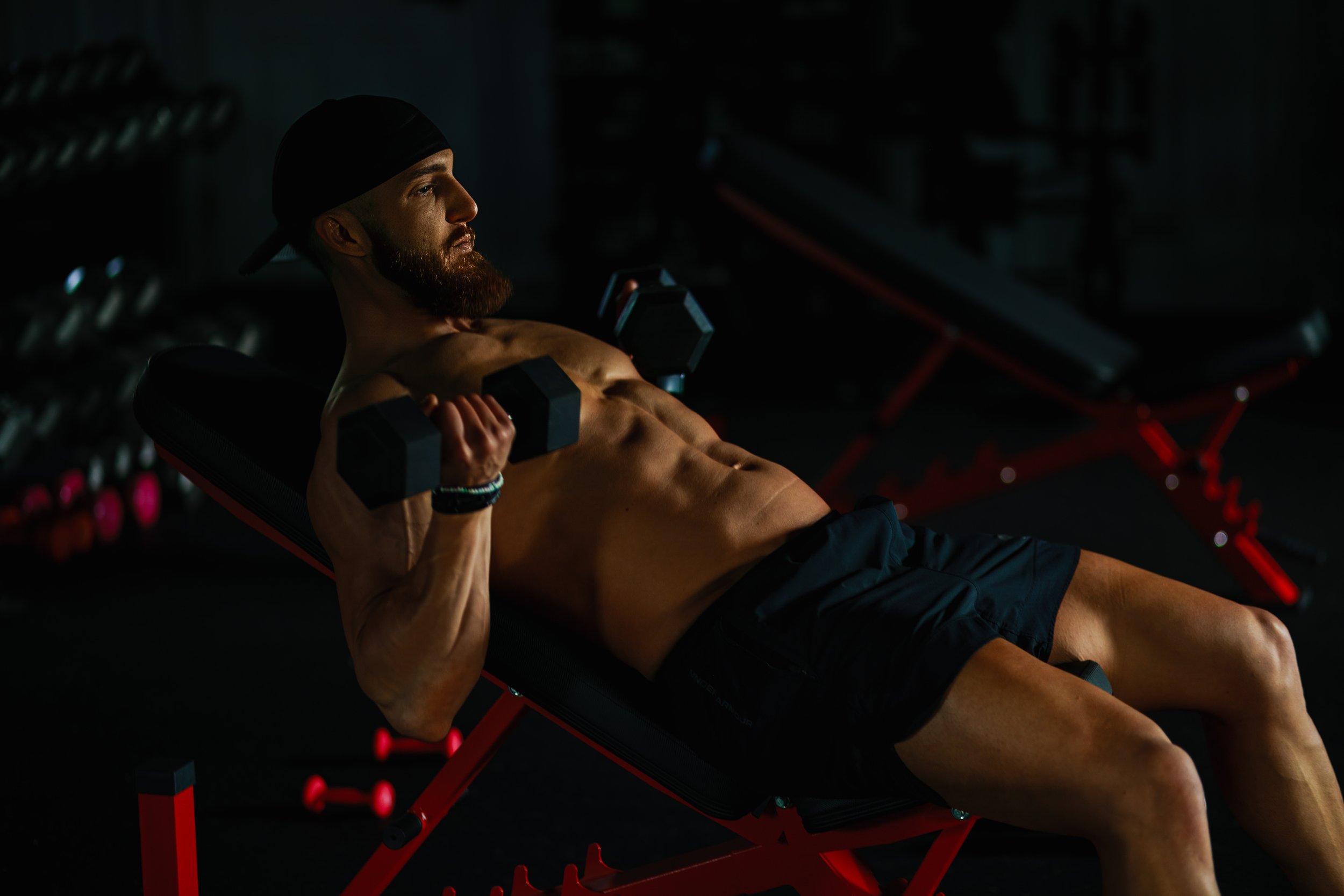 A muscular man with a beard and black cap lying on a workout bench, lifting a dumbbell in dark gym lighting.