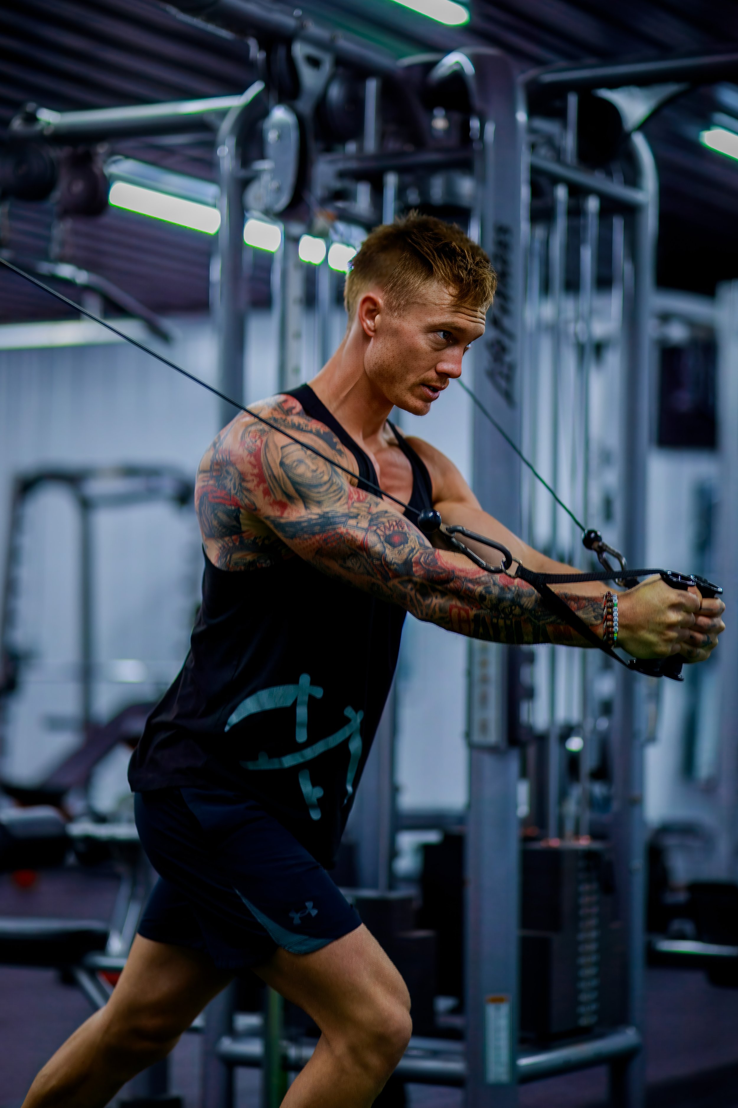 A young man with tattoos on his arms working out at a gym, using resistance bands tied to a machine.