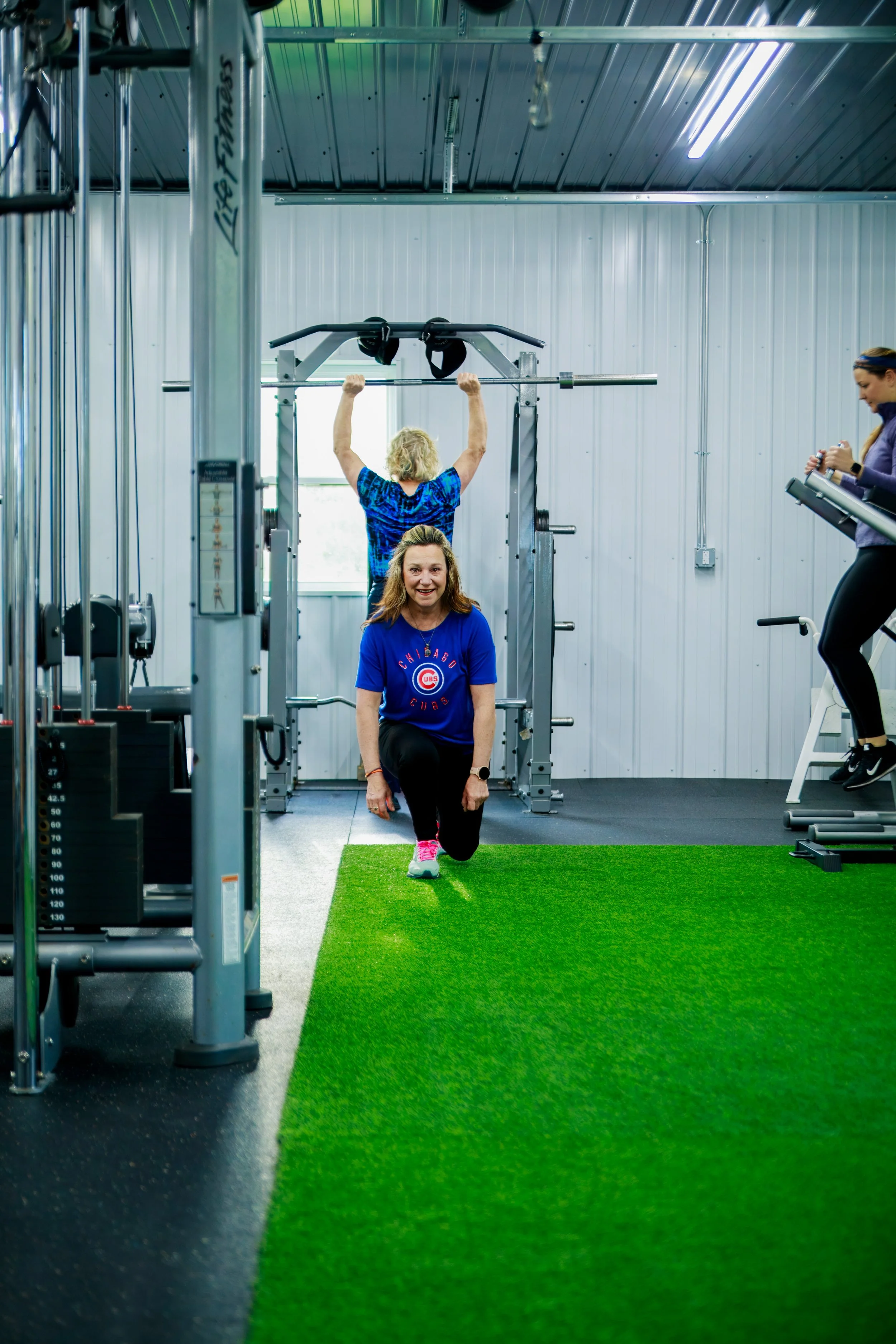 Three women exercising in a gym with weightlifting equipment. One woman is standing and smiling in the foreground, wearing a blue Chicago Cubs t-shirt. Another woman is lifting a barbell above her head in the background, while the third woman is on a