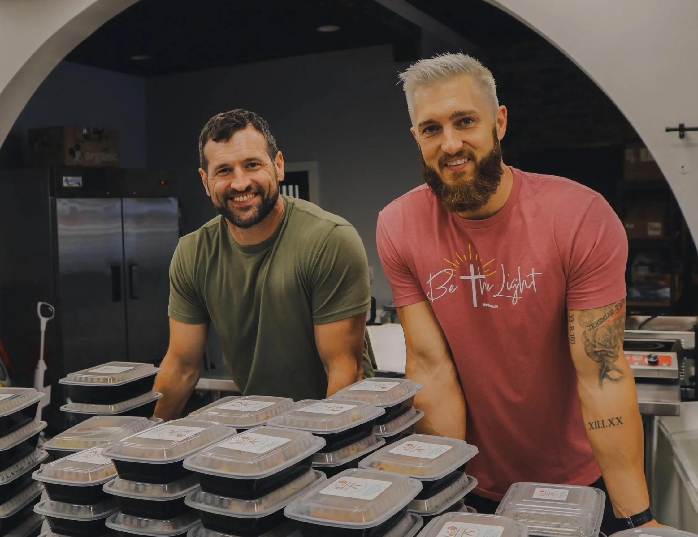 Two men smiling behind stacked meal containers in what appears to be a kitchen or food prep area.