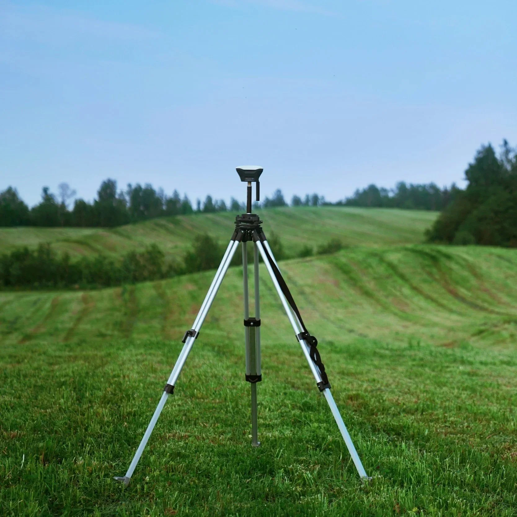A tripod stands on a grassy field with rolling hills and trees in the background under a clear blue sky.