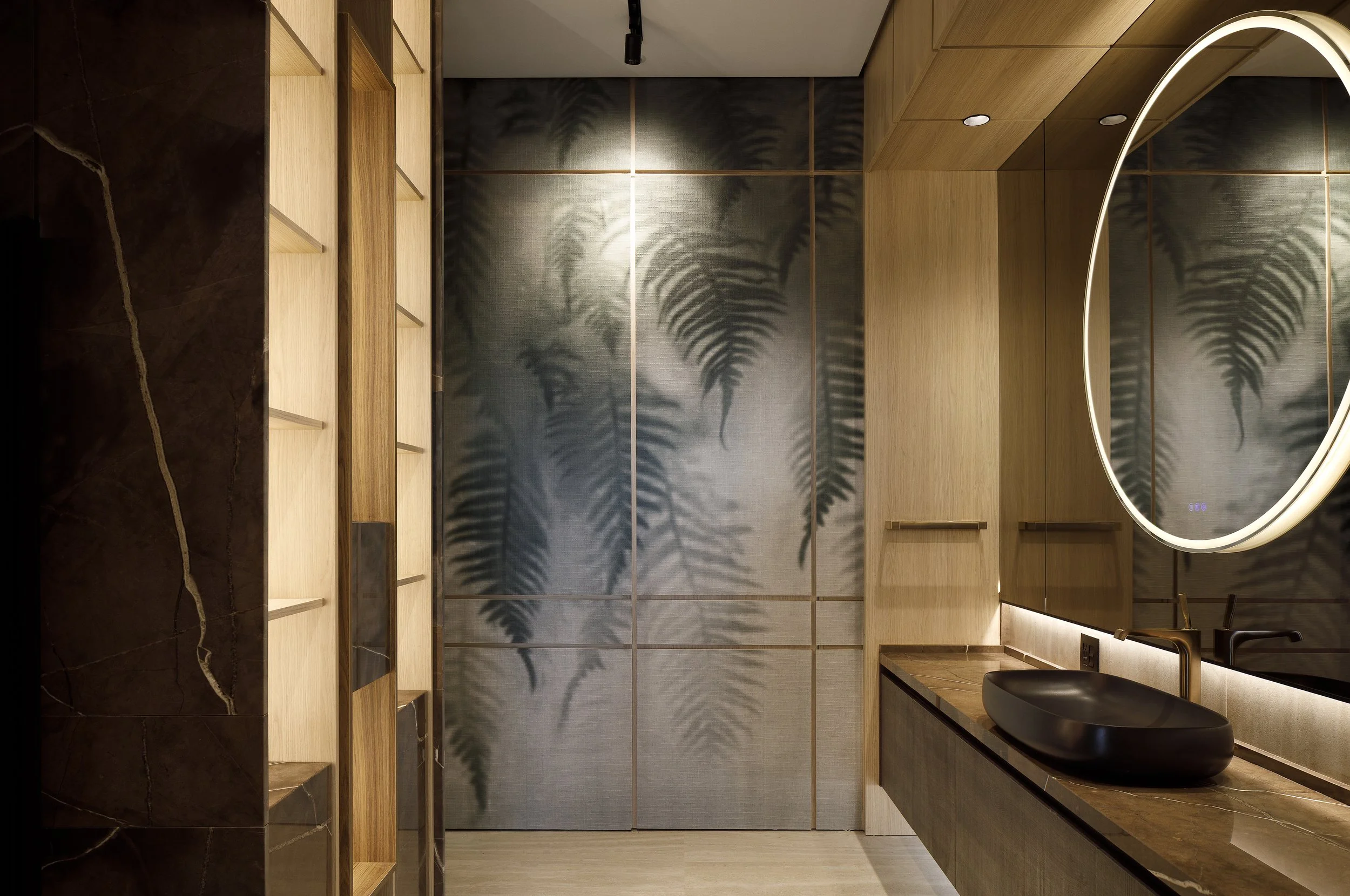 Baño con mural de motivos vegetales, espejo circular iluminado y lavabo sobre encimera oscura.