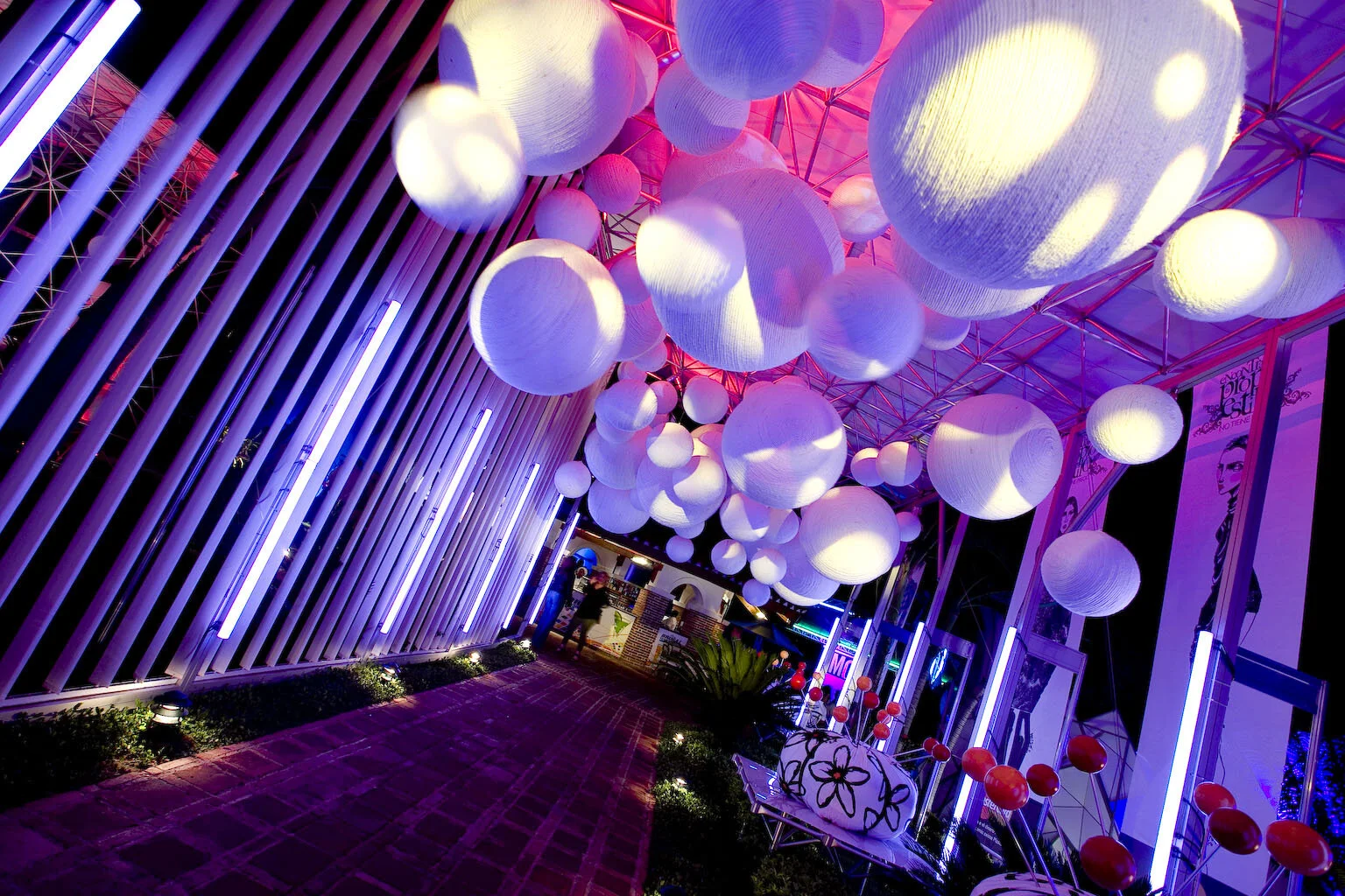 Decoración con globos de papel grandes blancos iluminados con luces de neón púrpuras y rosas, en un espacio moderno con paredes de varillas y posters en la pared, en medio de un ambiente nocturno y festivo.
