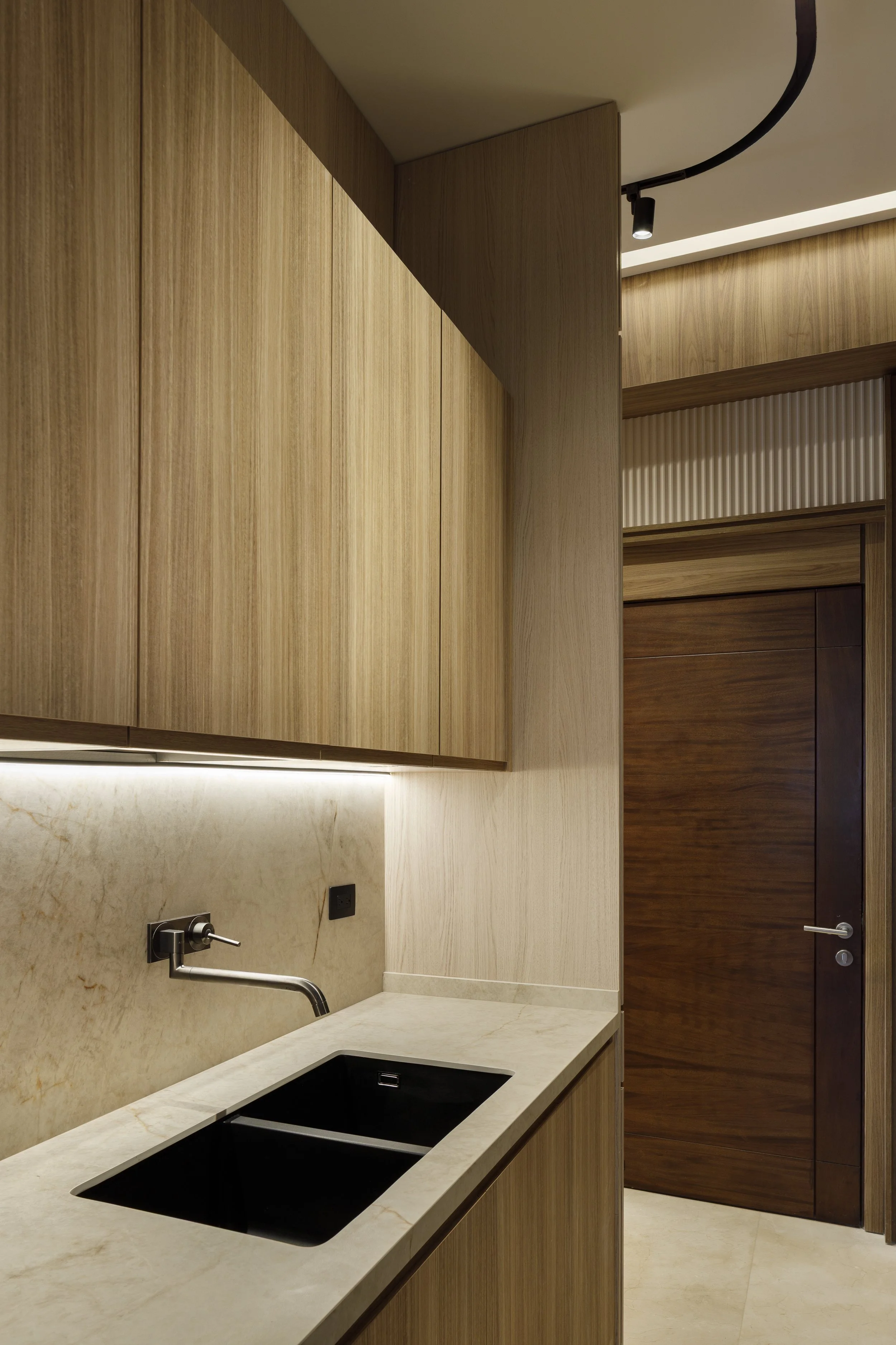 Una cocina moderna con gabinetes de madera clara, encimera de piedra, fregadero con grifo y pared de azulejos beige, con puertas de madera oscuras y una iluminación empotrada.