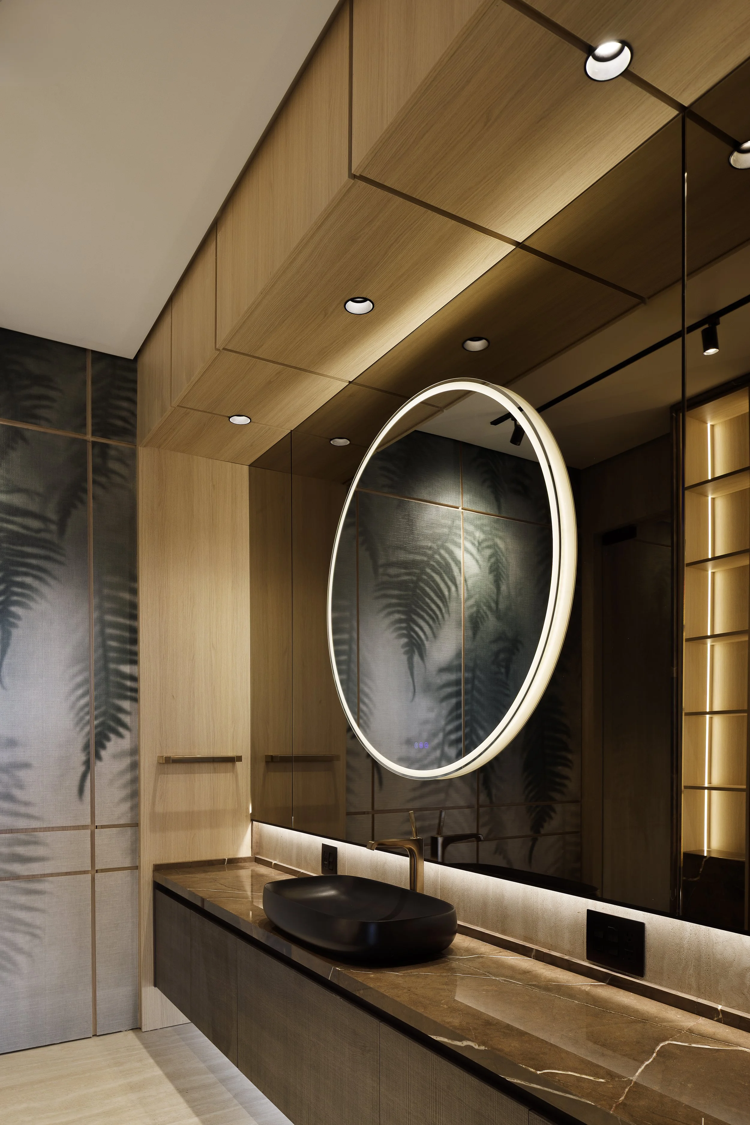 Lavabo negro sobre encimera veteada, con espejo circular iluminado y mobiliario de madera.