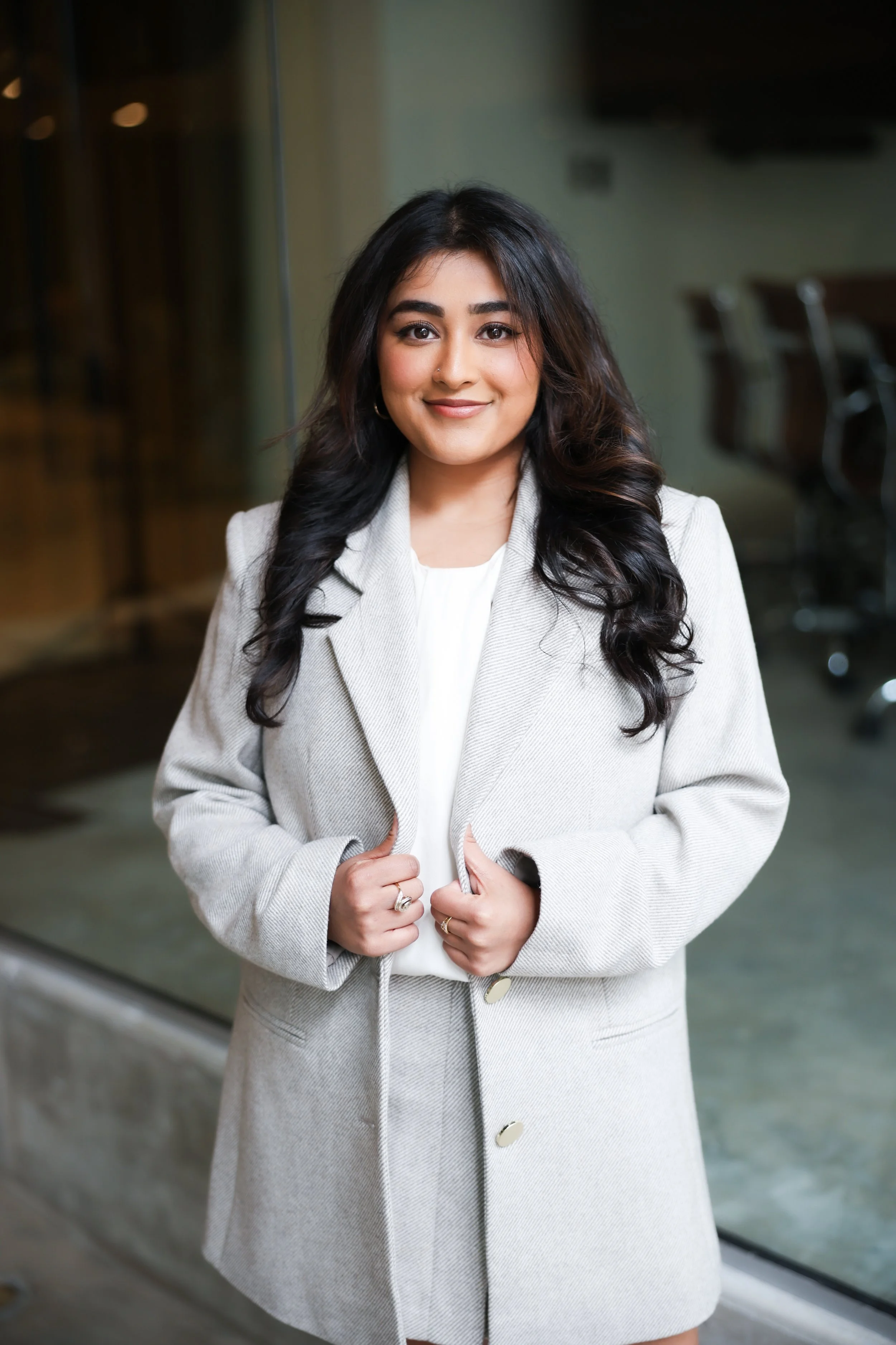 A young woman with long dark hair in loose curls, wearing a light gray blazer and skirt with a white blouse, smiling and leaning against a perforated metal railing in an indoor setting with wooden and glass elements.