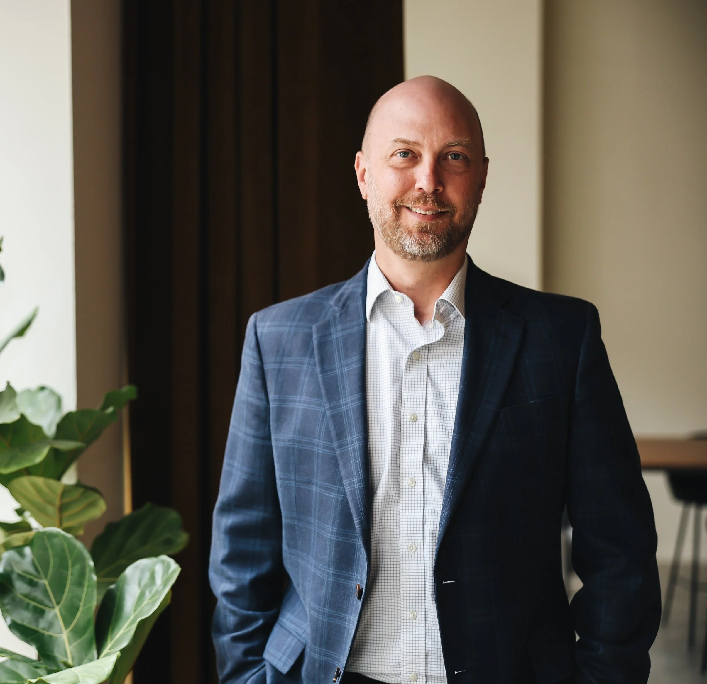 A smiling man in a checked white shirt and a dark blue plaid blazer standing indoors near a plant.