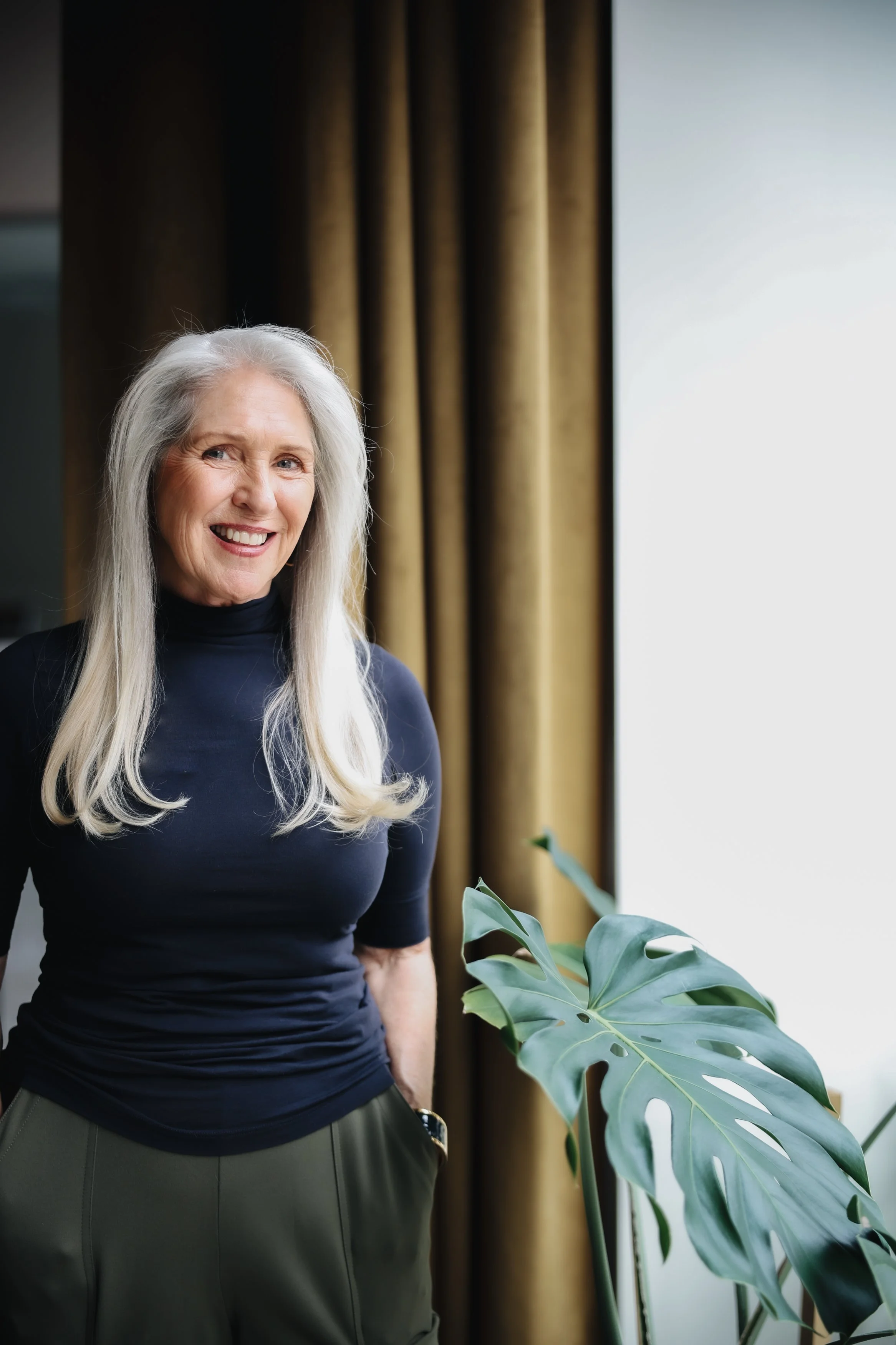 An older woman with long gray hair smiling, standing indoors near a large window with gold curtains, wearing a navy blue top and green pants, with a large green monstera plant nearby.