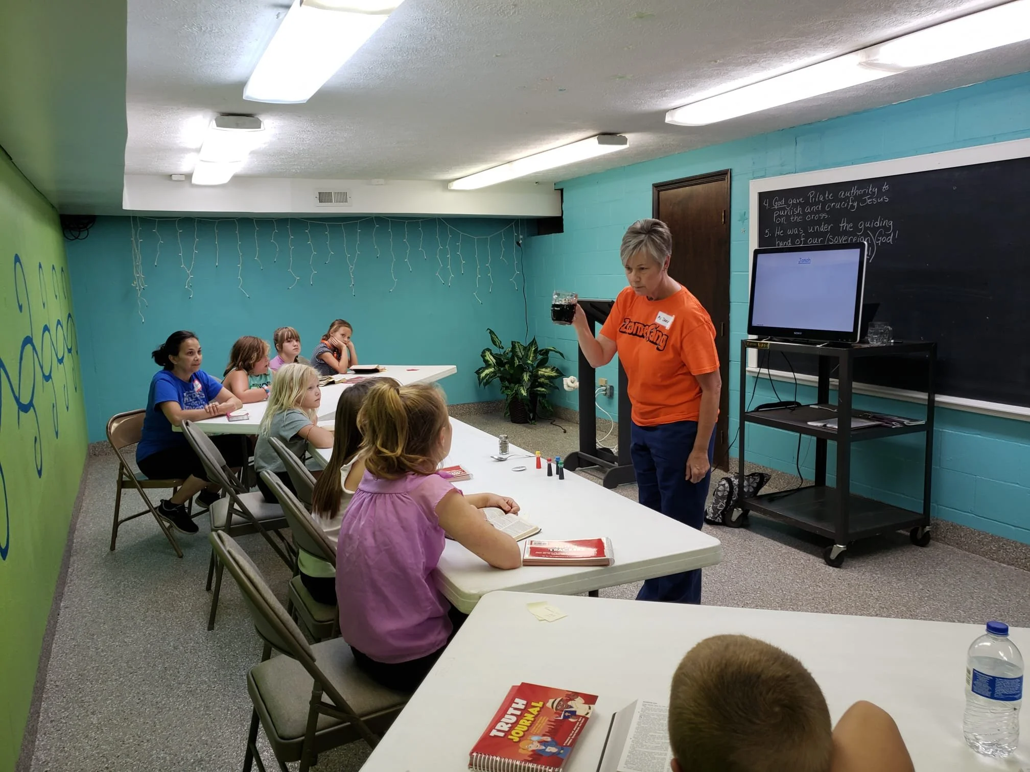 A classroom with young students seated at tables listening to a teacher who is standing at the front, holding a drink. The classroom has colorful walls, string lights hanging from the ceiling, and a blackboard with writing.
