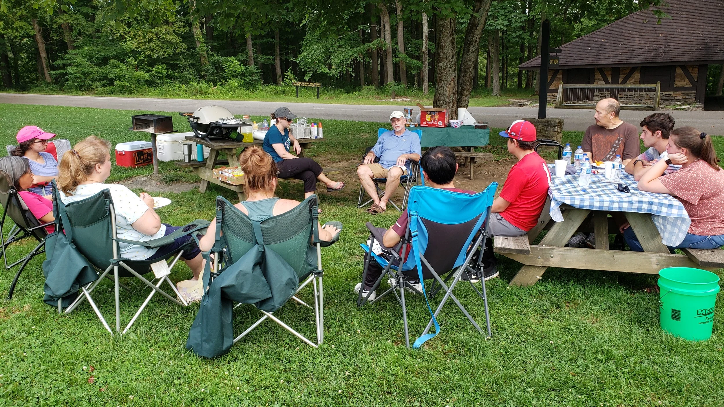 Group of people gathered outdoors for a picnic or barbecue, sitting on chairs and benches under trees with a picnic table and barbecue grill nearby, surrounded by greenery.