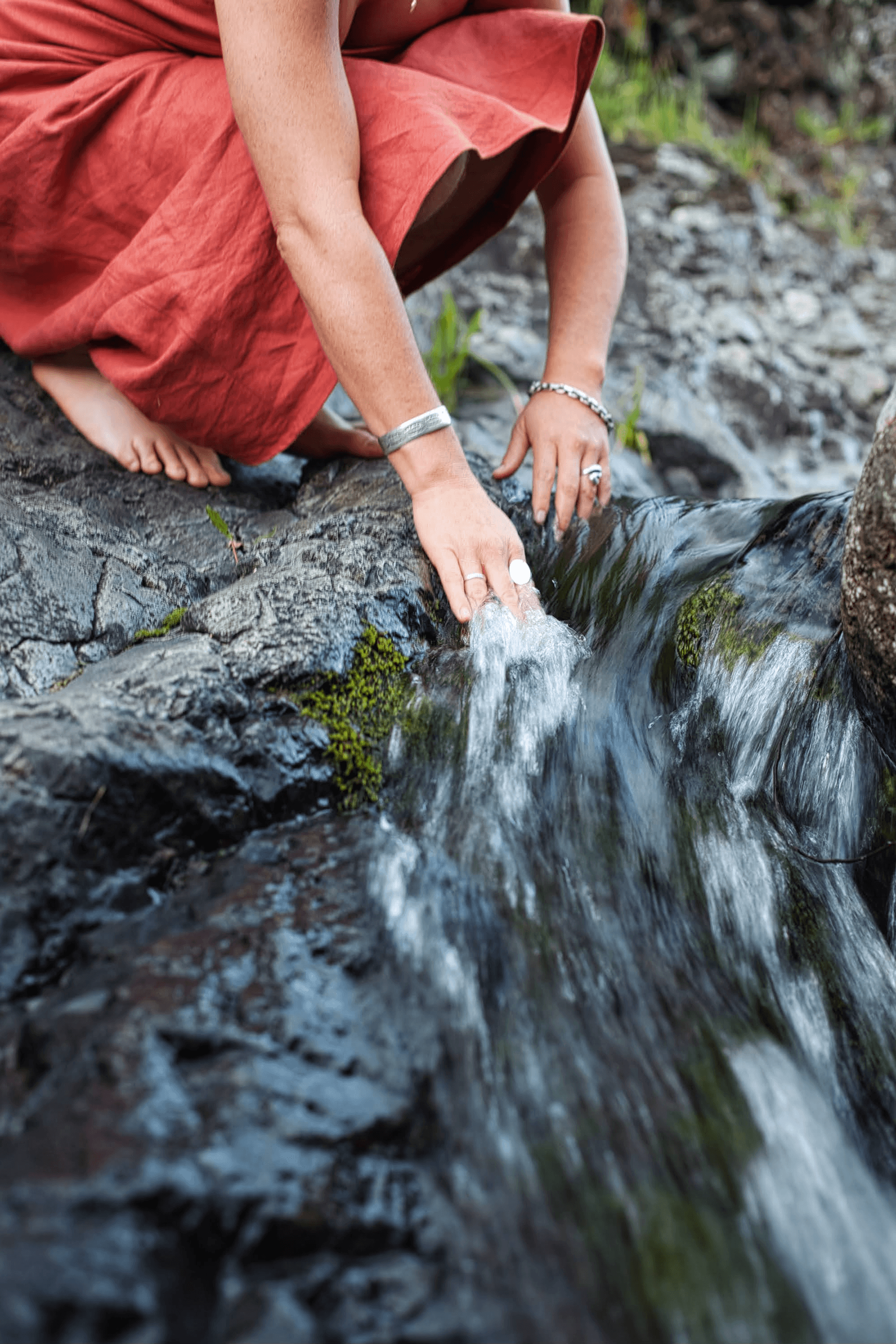 A person crouching down by a small creek in a natural outdoor setting, wearing a red dress and silver jewelry, touching the flowing water with their hands and bare feet visible.