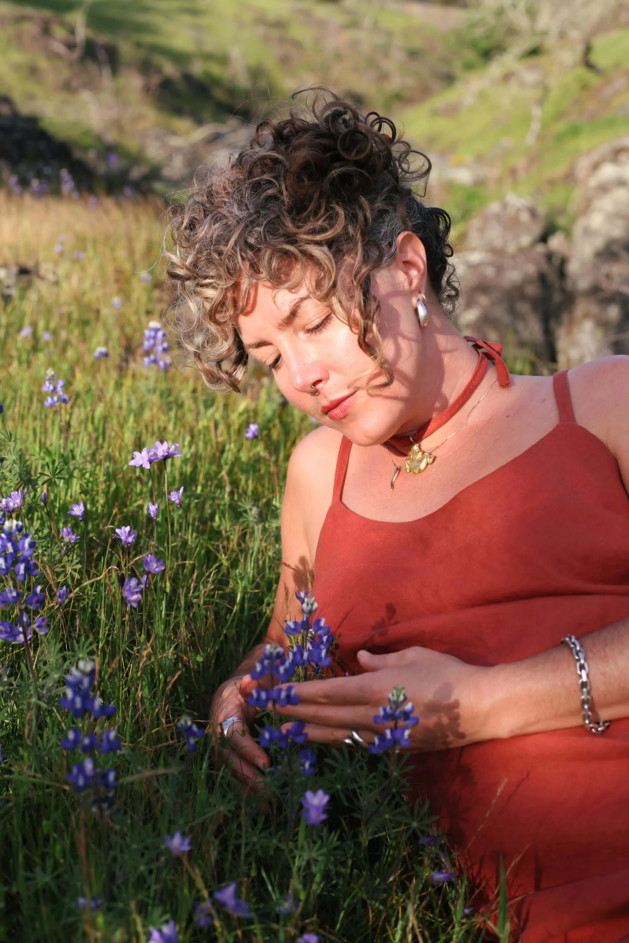 A woman with short curly hair and earrings, lying in a field of purple flowers with a hillside in the background, wearing a rust-colored tank top.