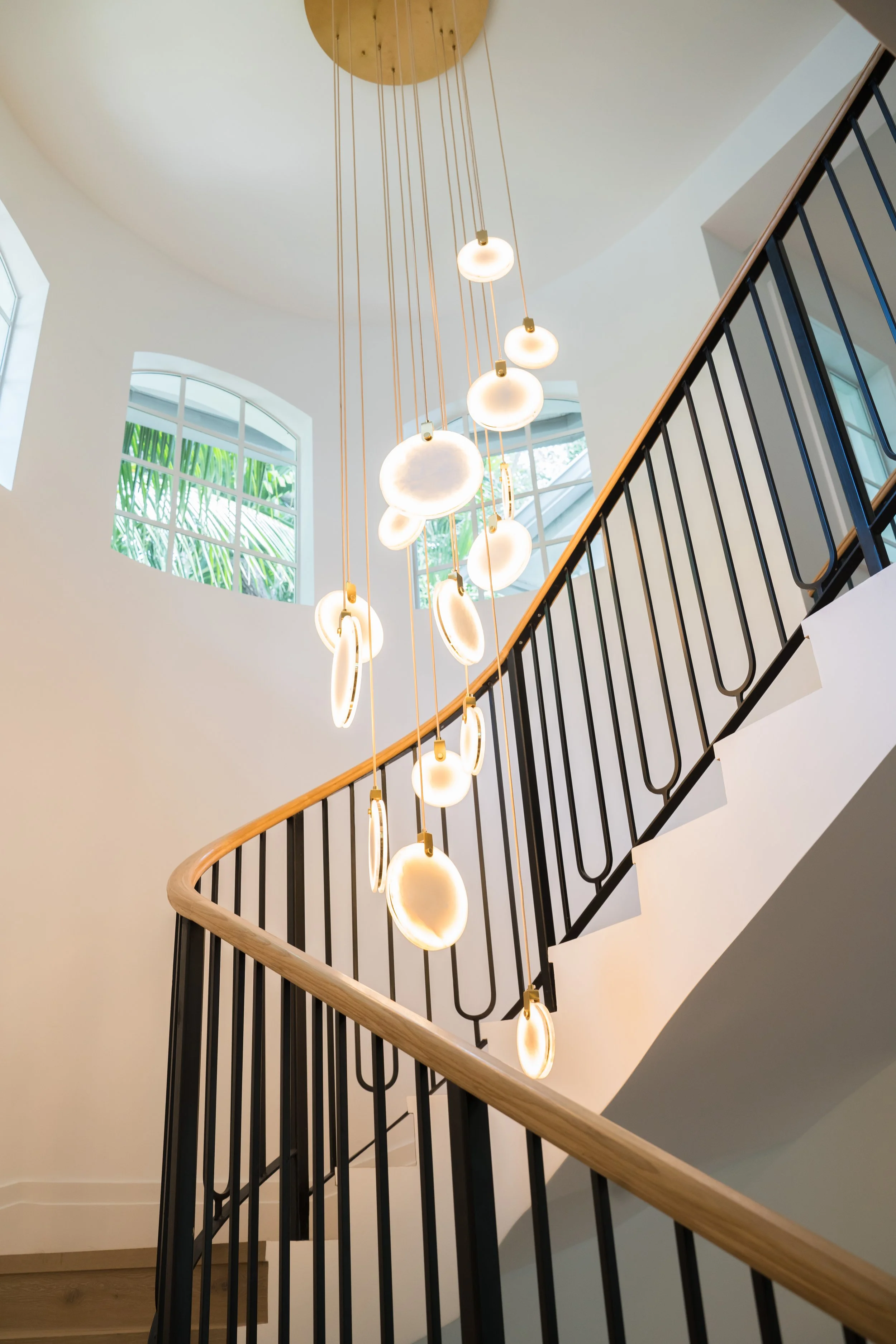 Modern staircase with a wooden handrail and black metal balusters, illuminated pendant lights hanging from the ceiling, and large windows letting in natural light.