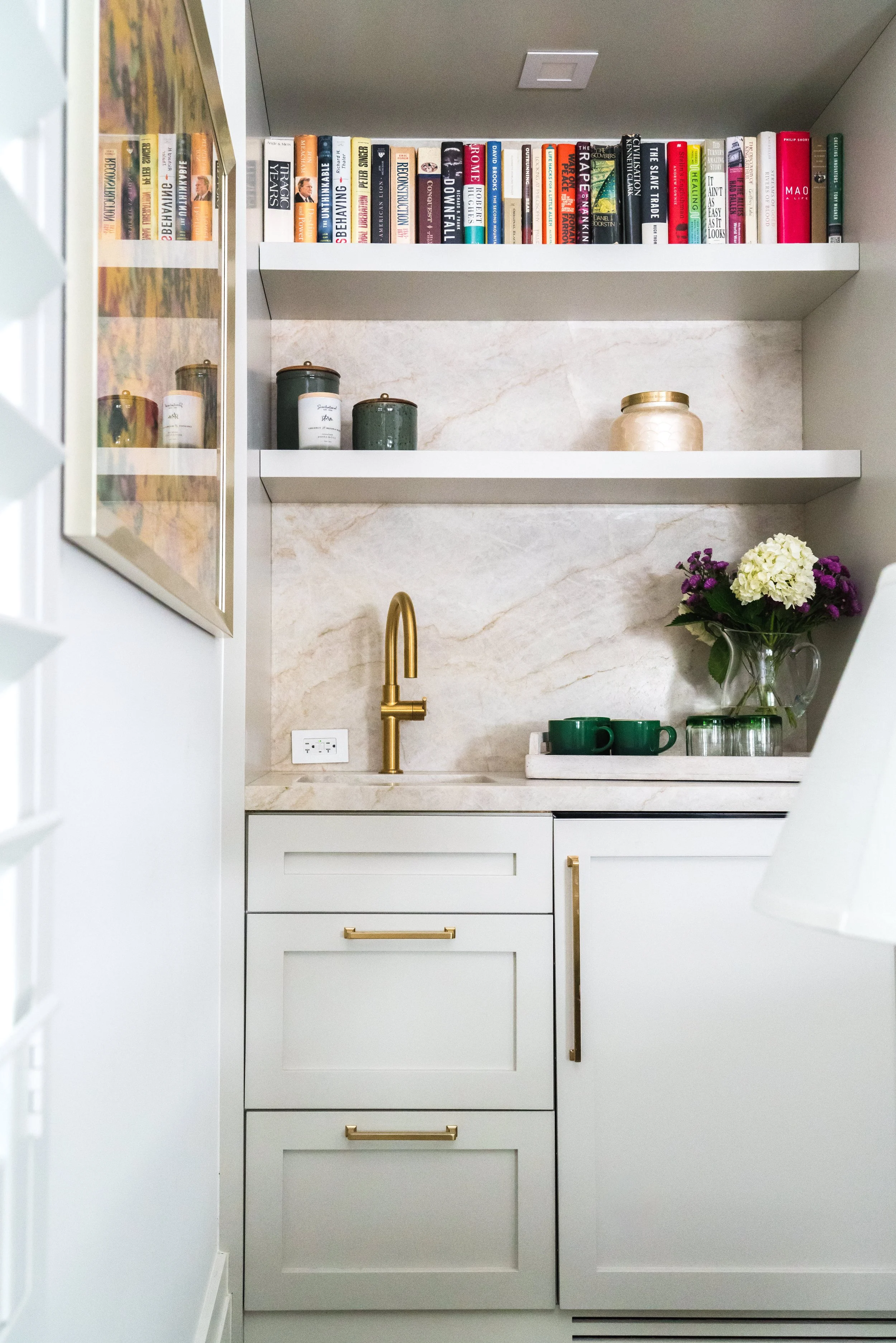 A kitchen with white cabinets, gold hardware, a gold faucet, and a marble backsplash. Open shelves hold green jars and a vase with white and purple flowers. A power outlet is visible on the wall.