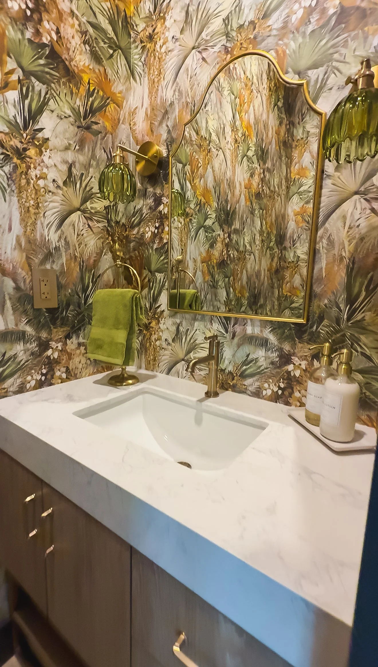 Bathroom sink with marble countertop, gold fixtures, and a gold-framed mirror on a tropical patterned wallpaper background. There are green towels and soap dispensers on the counter.
