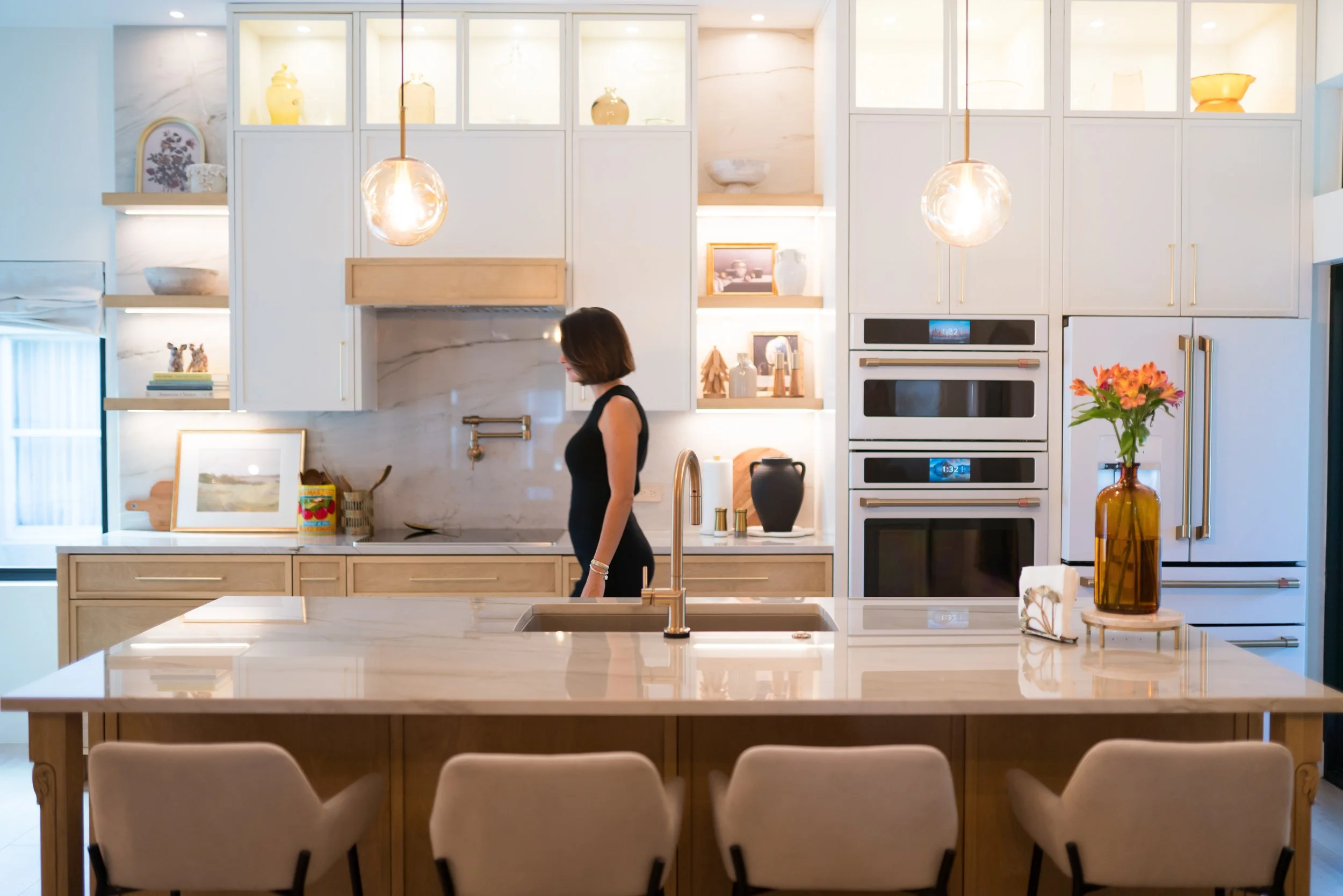 Modern kitchen featuring a large white marble island with seating, light wood lower cabinets, white upper cabinets, a built-in double oven, decorative vases, and a woman standing near the sink, with warm lighting and minimalist decor.
