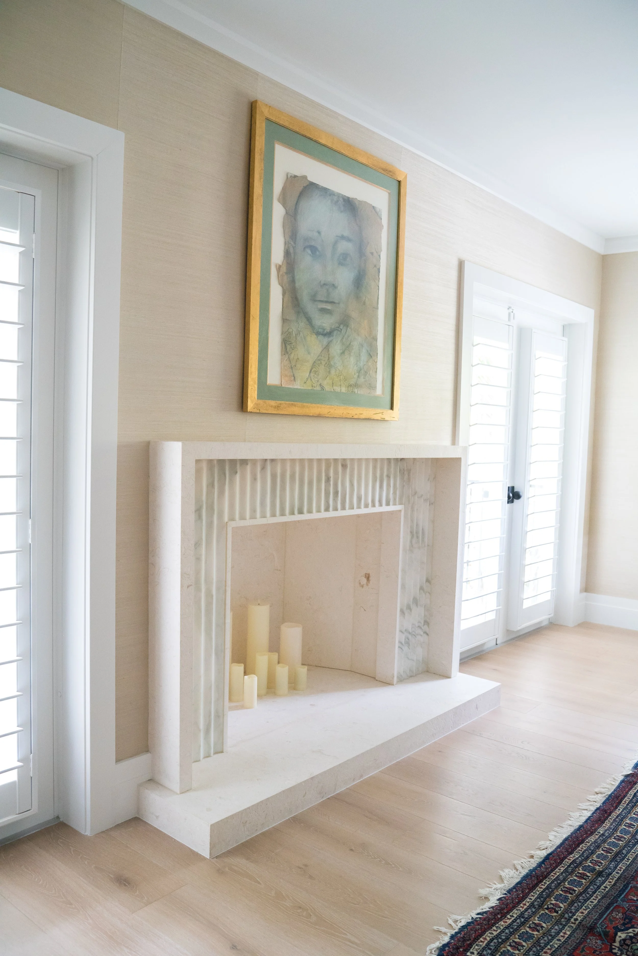 A living room with a white fireplace, light wooden flooring, and a colorful Persian rug. There is a framed watercolor portrait of a woman hanging on the wall above the fireplace. The room has two large windows with white shutters.