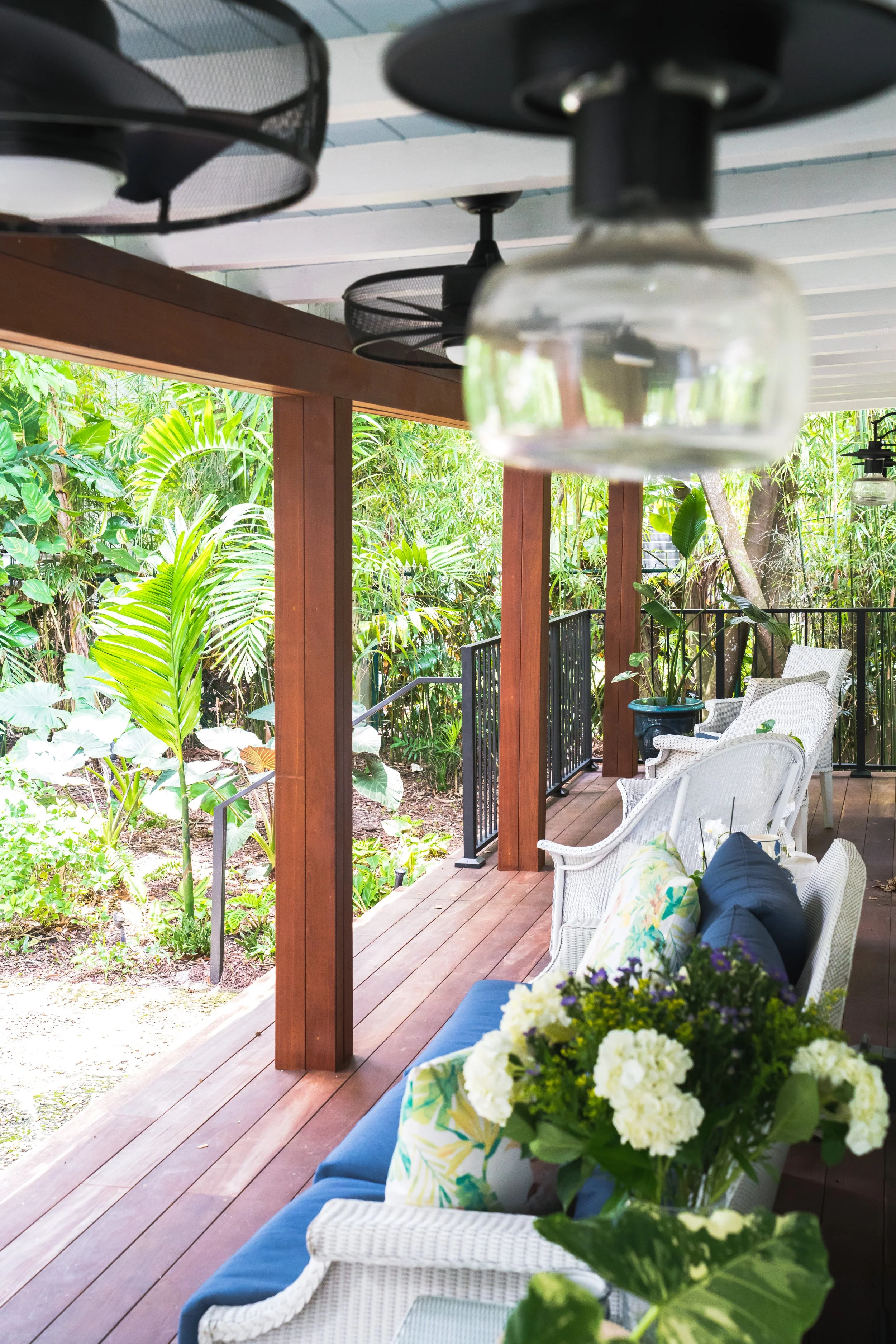 A porch with wooden flooring, white wicker chairs with cushions, a potted plant, and hanging ceiling lights, surrounded by lush greenery.