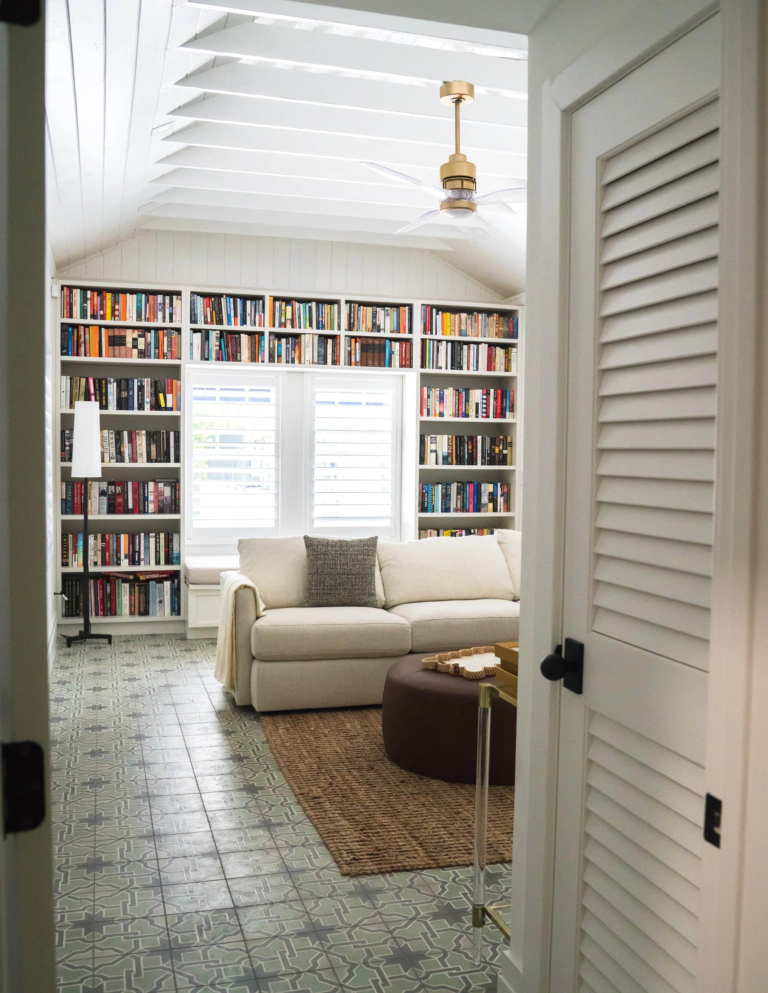 A cozy living room with a white sofa, brown ottoman, and a small side table with decor, viewed through a partially open louvered door. The room has tall bookshelves filled with books, a window with closed blinds, a ceiling fan, and patterned tile flooring.