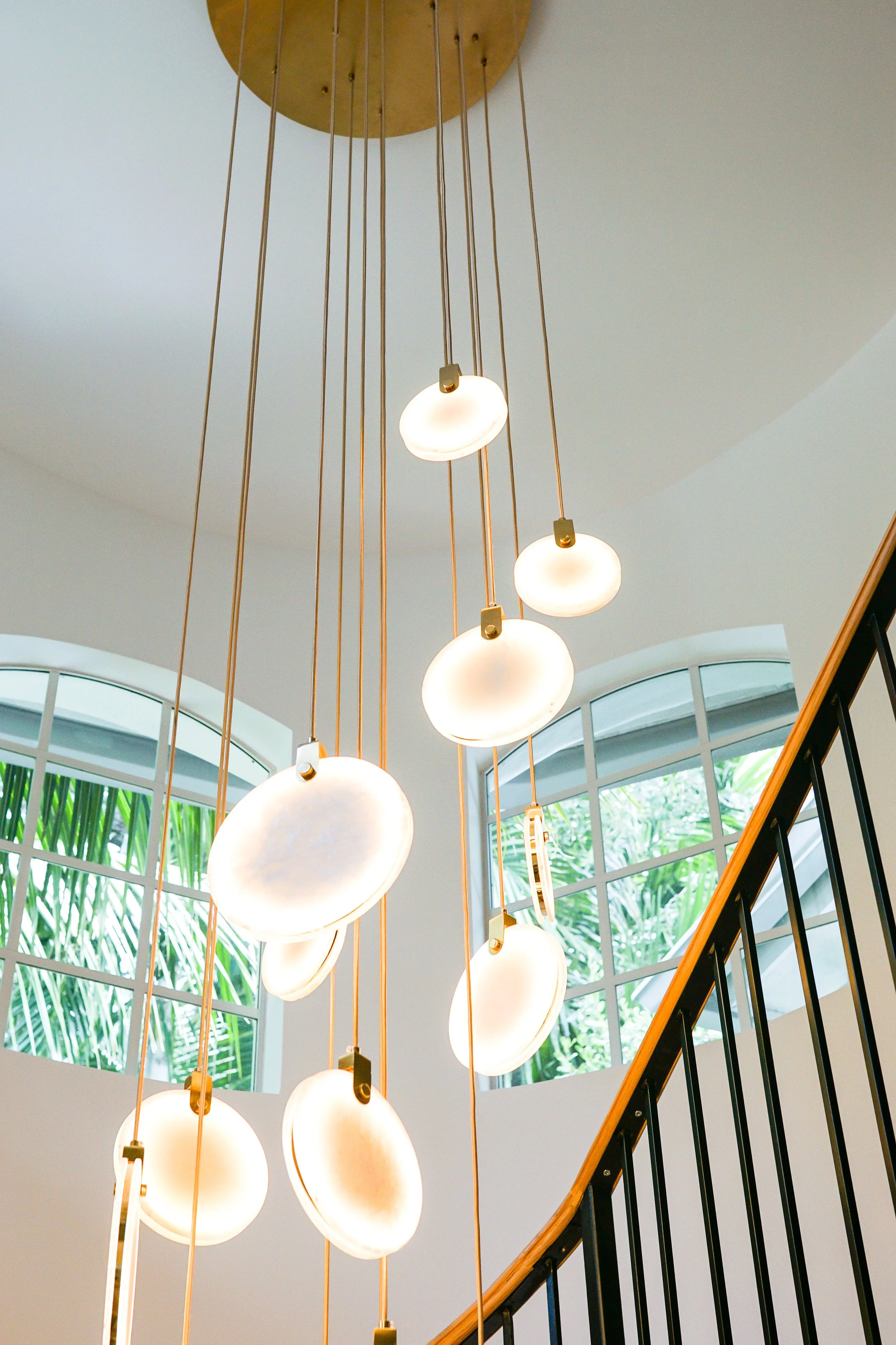 A modern chandelier with multiple frosted glass globes hanging from gold-colored rods, positioned above a staircase with a black metal railing and a curved wooden handrail, in front of large arched windows with greenery outside.