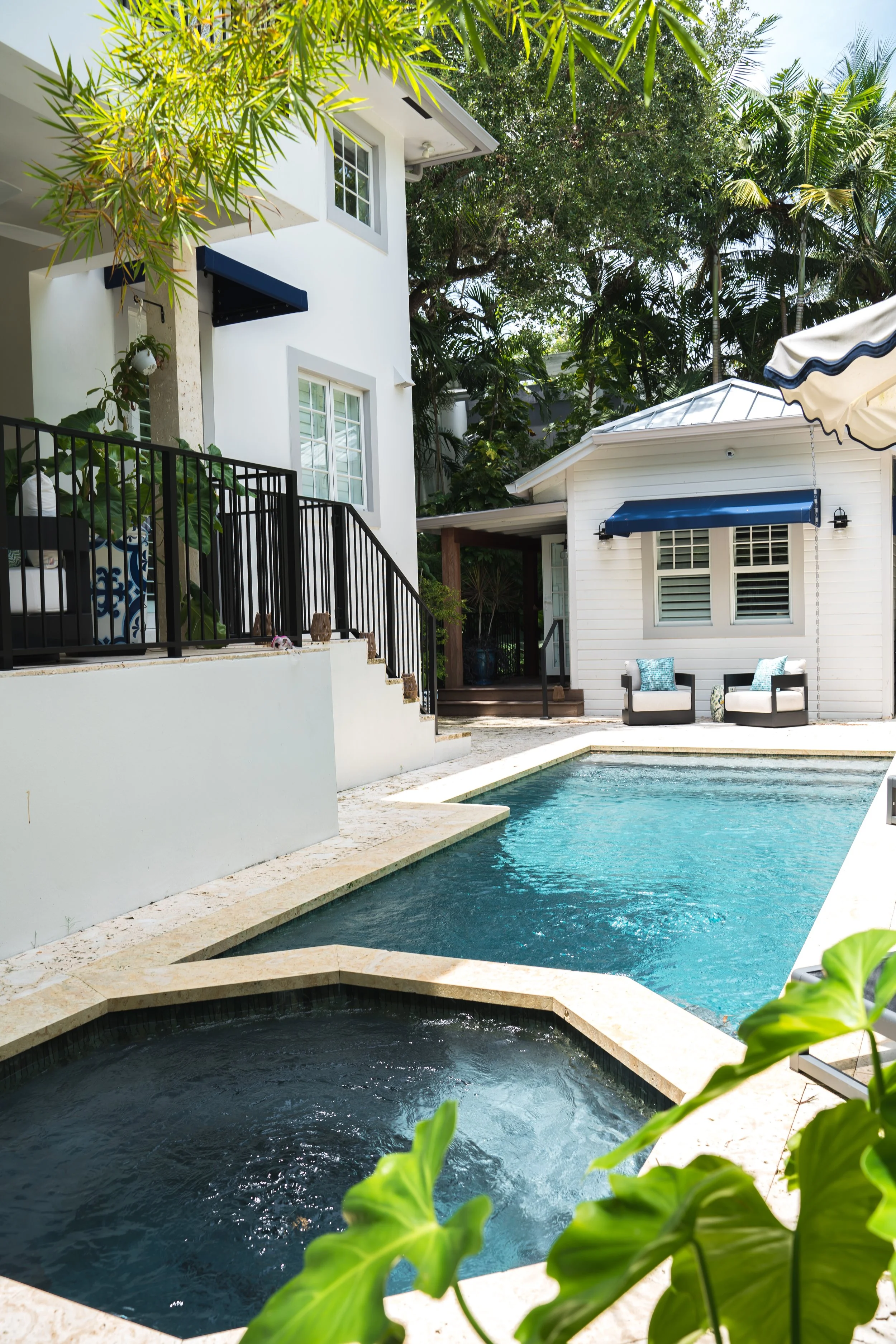 A backyard pool area with a hot tub in the foreground, a kidney-shaped swimming pool in the middle, and two chairs with patterned cushions near a small white building with blue awnings and a seating area with a patio umbrella. Green trees and plants surround the area.