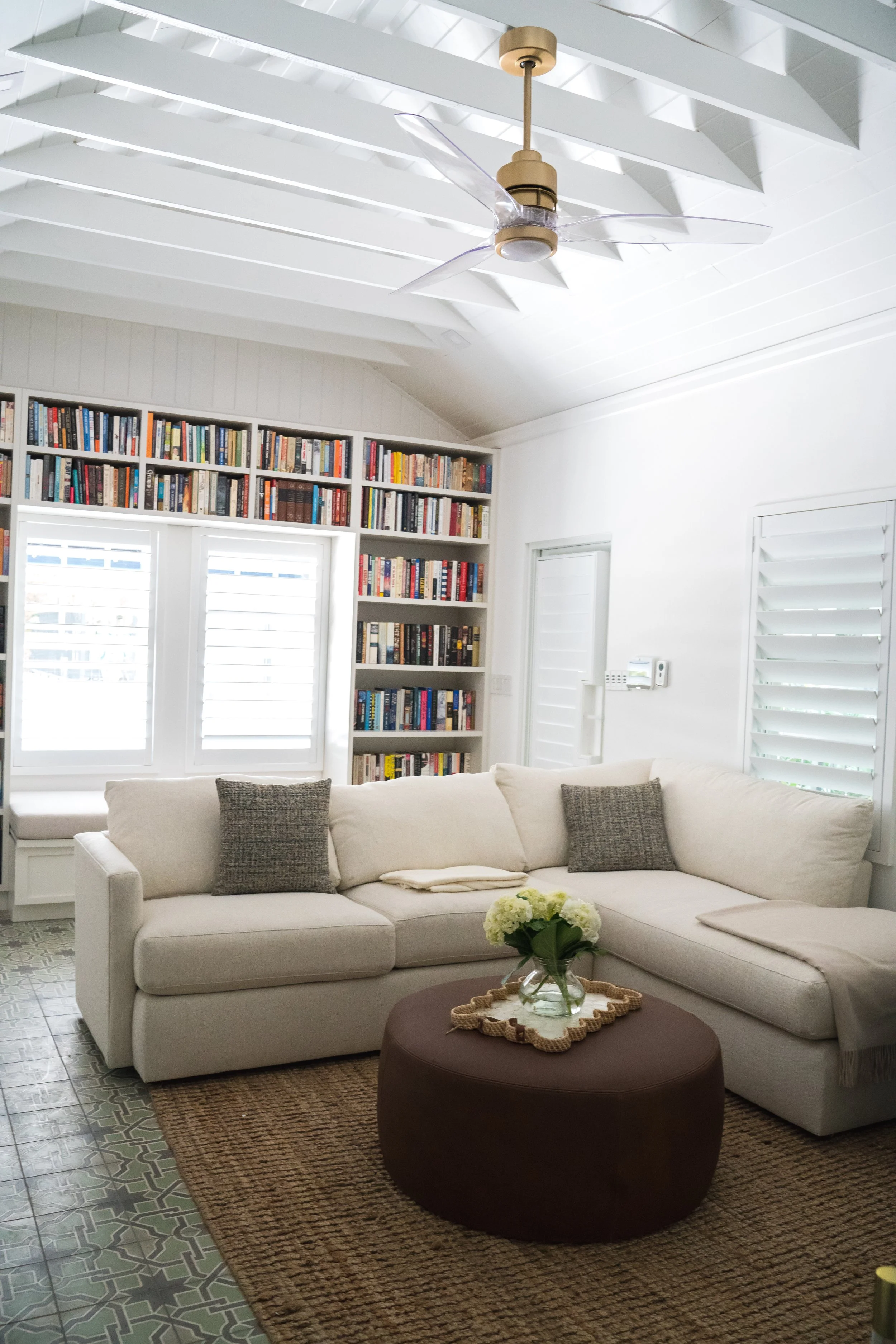 A cozy living room with a white sectional sofa, gray cushions, a glass vase with white flowers on a round brown ottoman, a woven area rug, white built-in shelves filled with books, white plantation shutters, and a ceiling fan with light fixture under a vaulted ceiling with white exposed beams.