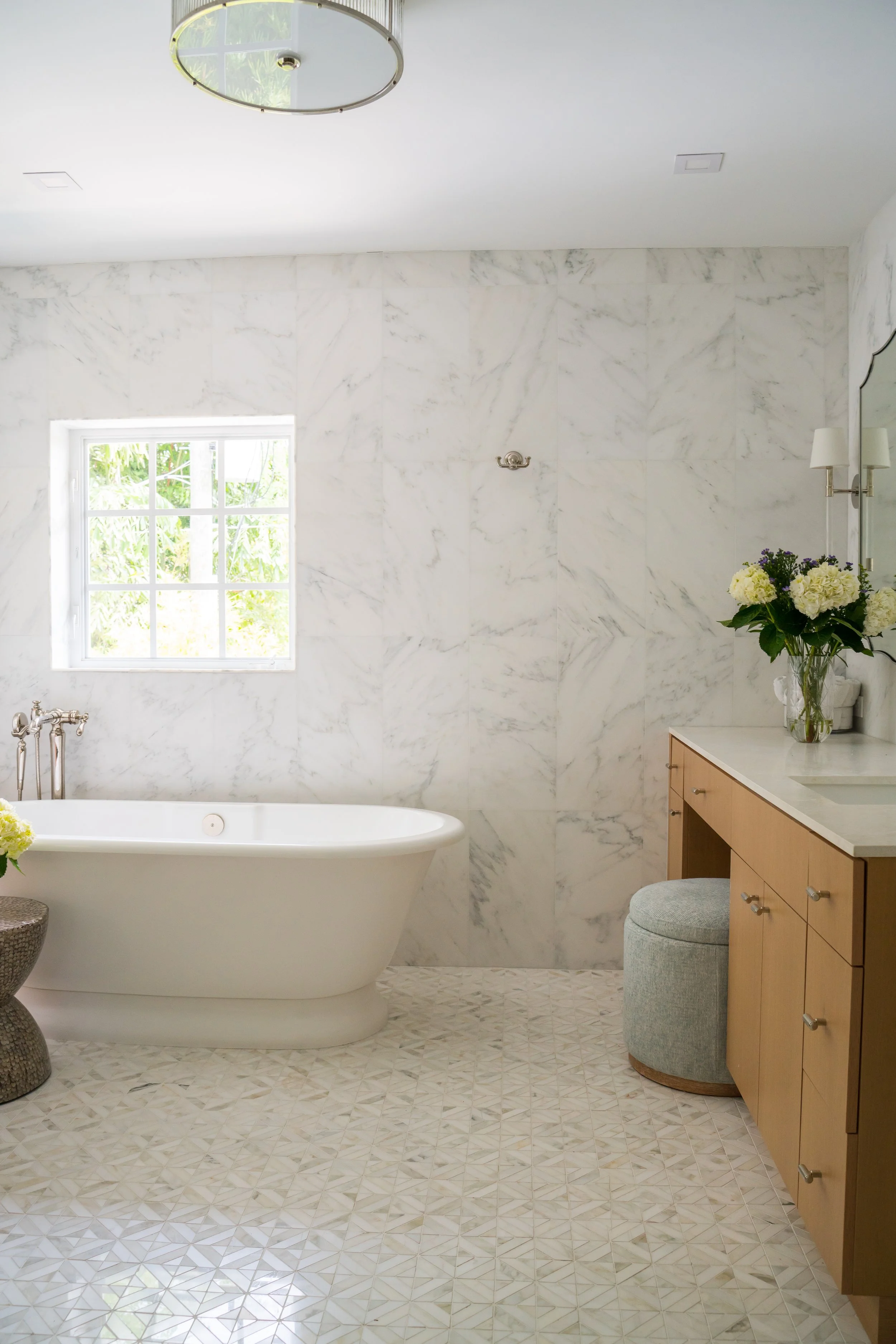 Bright bathroom with white marble walls, a freestanding bathtub, window with greenery outside, floral arrangements, and beige cabinetry.