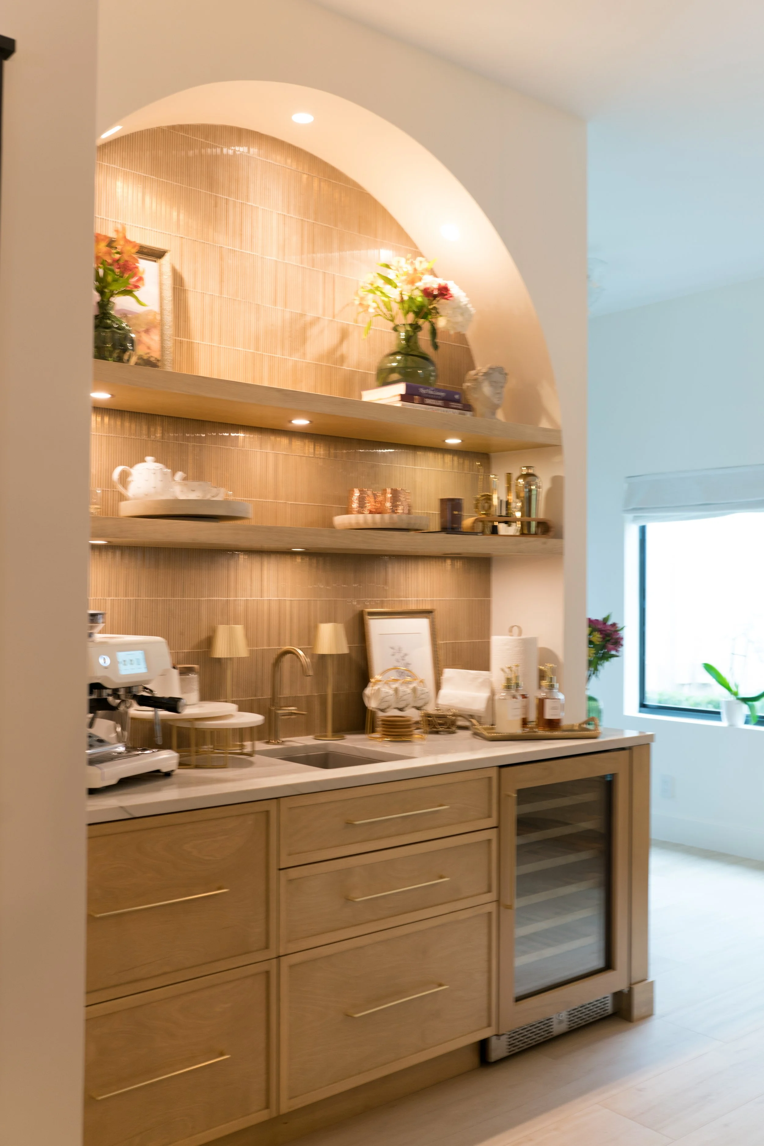 A kitchen area with a wooden cabinet, a coffee machine on the countertop, and open shelving above displaying decorative items, books, and a vase with flowers. A window is visible on the right side.
