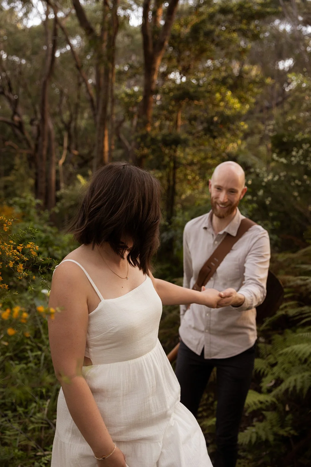 A man and a woman, both dressed in light-colored clothing, are embracing and smiling at each other on the beach during sunset. The man is lifting the woman off the sand, and they are near the shoreline with gentle waves in the background.