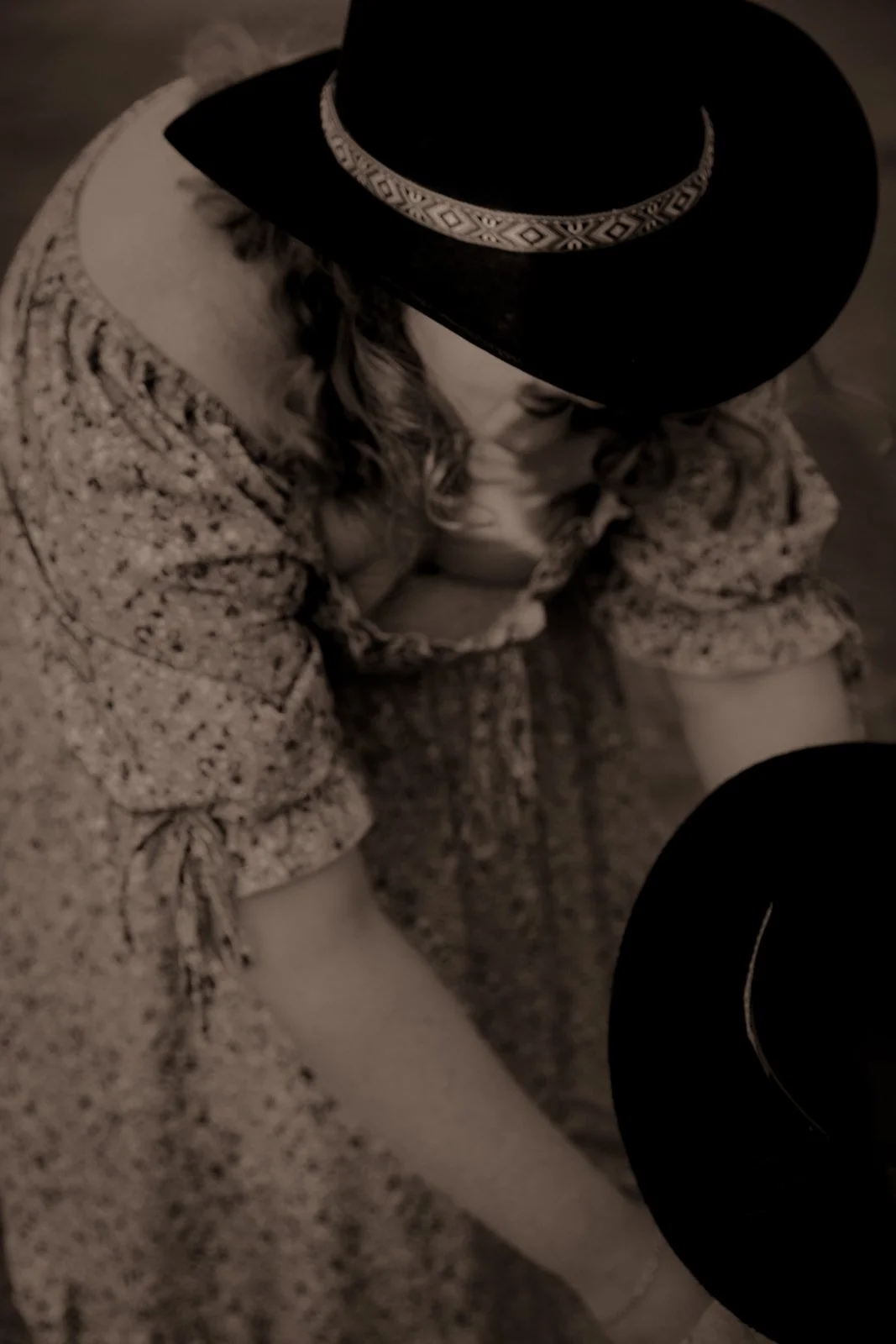 A person wearing a cowboy hat with decorative band, a patterned shirt, and a jacket with bow details on the sleeves, sitting with arms crossed, captured from a top-down perspective in black and white.