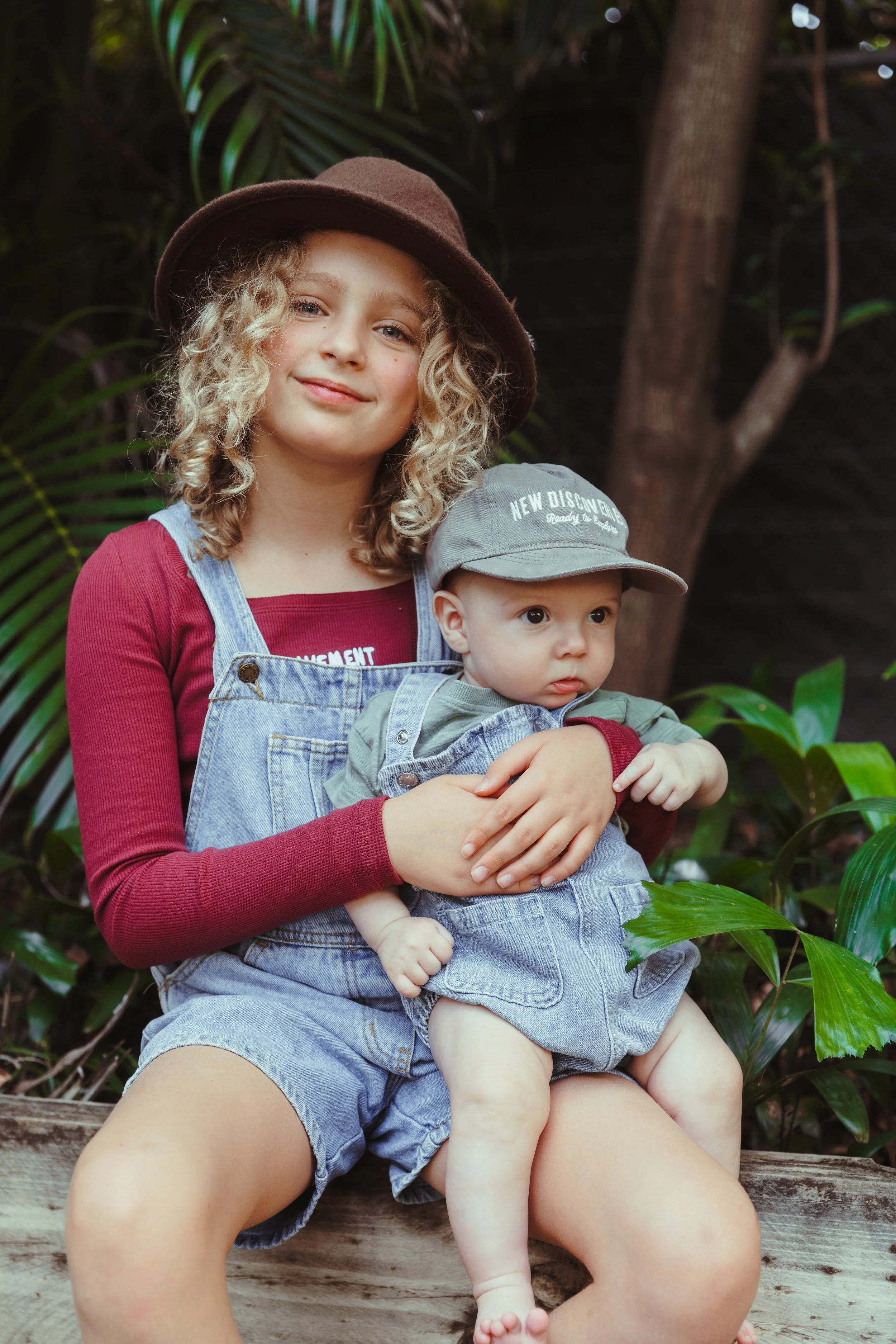 A girl with curly blonde hair wearing a brown hat, a maroon long sleeve shirt, and denim overalls sitting outdoors with a baby on her lap. The baby is wearing a gray cap with text, green shirt, and denim shorts, sitting on the girl's lap surrounded b